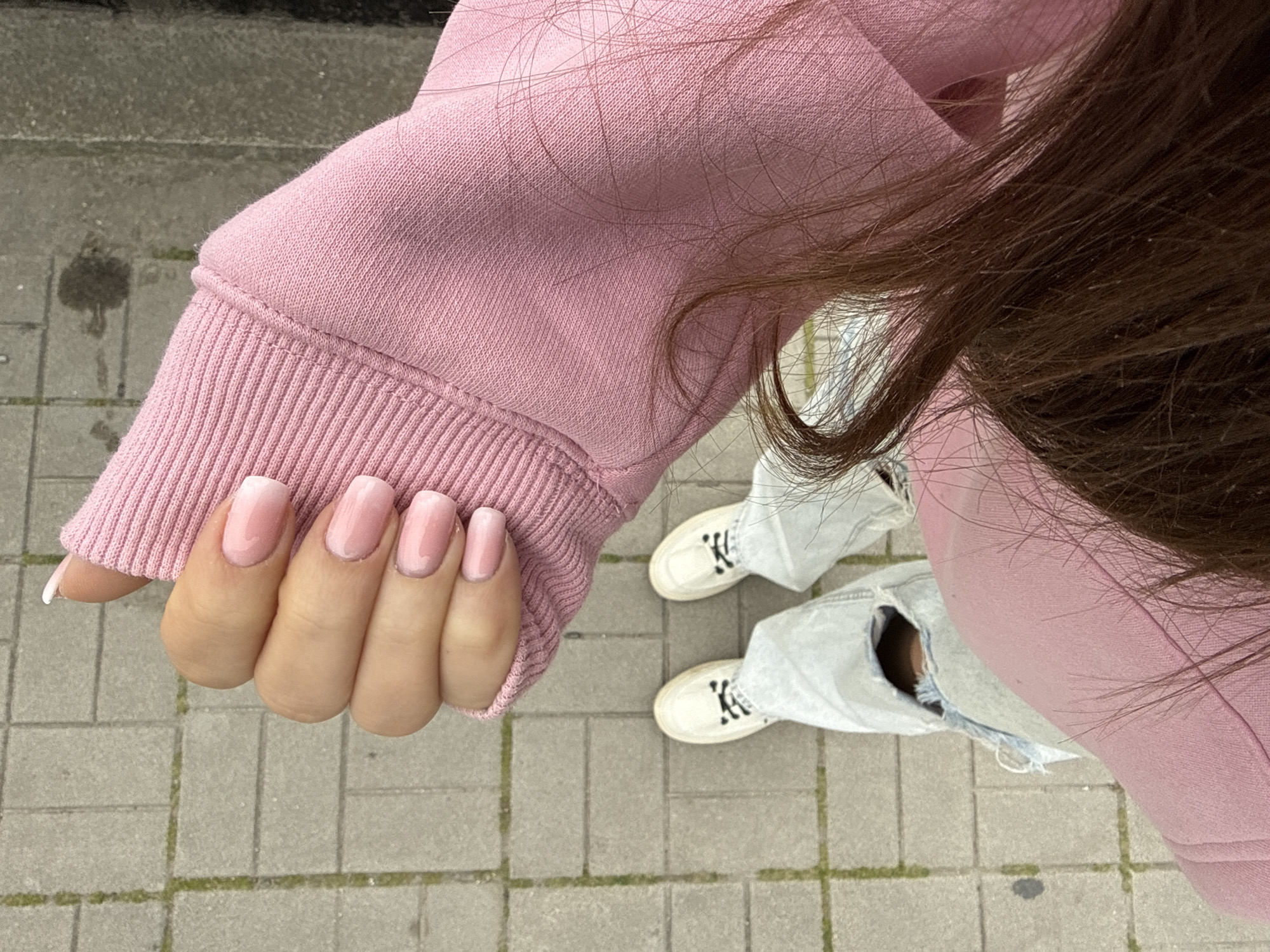 The image is a first-person perspective shot taken outdoors. The subject is wearing a pink hoodie and distressed light-wash jeans with holes at the knees, paired with white sneakers with black accents. The person's hand, with pink ombre manicured nails, is positioned in the foreground, resting on the hoodie sleeve. The background features a paved pedestrian area, possibly a sidewalk, with grey rectangular stones. The hair of the subject falls in the upper right portion of the image.
There is no information available in this image that indicates that the photograph was taken in Plovdiv, Bulgaria.