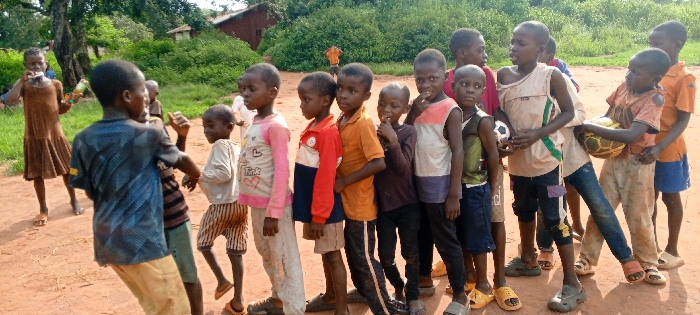 The image displays approximately fifteen children outdoors on a reddish-brown dirt surface in the city of Ngalula, Democratic Republic of the Congo. The children, predominantly boys, appear to be between the ages of approximately 5 and 12 years. They are dressed in various casual clothing, including t-shirts, tank tops, shorts, and trousers; some garments exhibit signs of wear or dirt. Footwear consists primarily of sandals or flip-flops, with some children appearing barefoot.
In the center-right of the frame, one boy in a light-colored tank top holds a black and white soccer ball. To his immediate right, another boy in an orange, torn t-shirt holds a worn or deflated yellow and black patterned soccer ball with both hands. On the far left, a girl in a brown dress brings a small bottle with a green cap to her mouth. The children are positioned in a loose group, with several forming a somewhat linear arrangement in the mid-ground. Their gazes are varied, with some looking towards the viewer and others looking elsewhere. The background consists of dense green foliage, trees, and indistinct structures partially visible through the vegetation on the left side of the frame. The scene is illuminated by natural daylight.