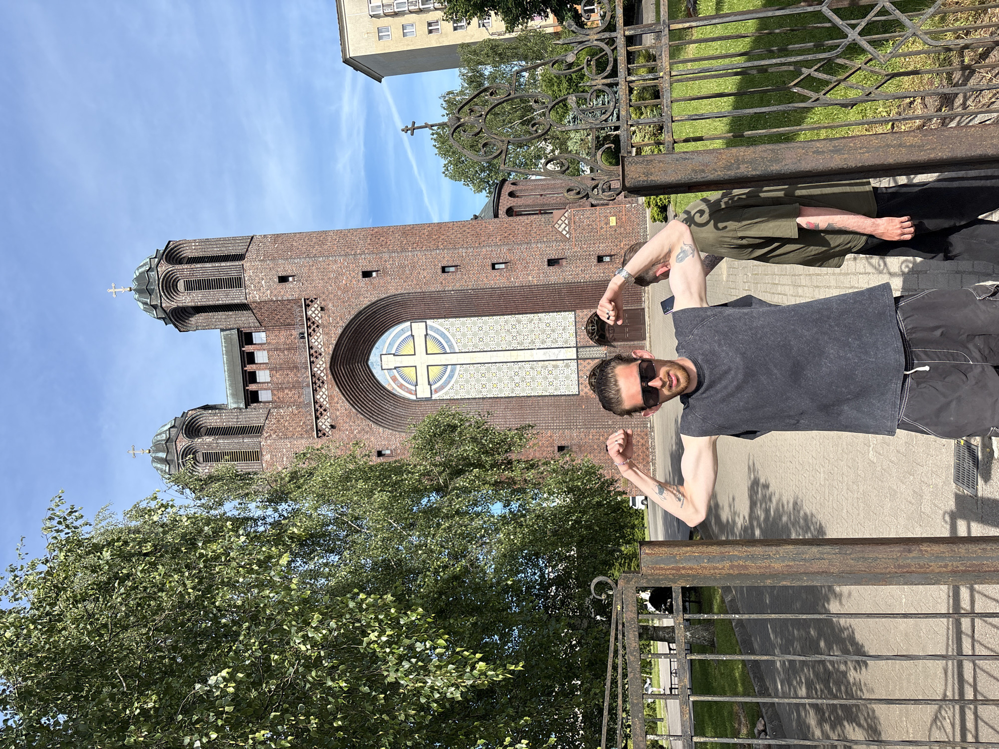 An outdoor, eye-level, medium shot shows a man flexing his biceps in front of a large brick church in Kaliningrad, Russia. The church features two towers with rounded tops, one with a cross and intricate arched windows. The man, wearing sunglasses, a dark sleeveless shirt, and shorts, stands by a rusty metal gate. A second person, wearing a green shirt, stands behind him to the right. The sun is shining, casting shadows on the paved ground and grass. The background includes trees and a tall apartment building.
