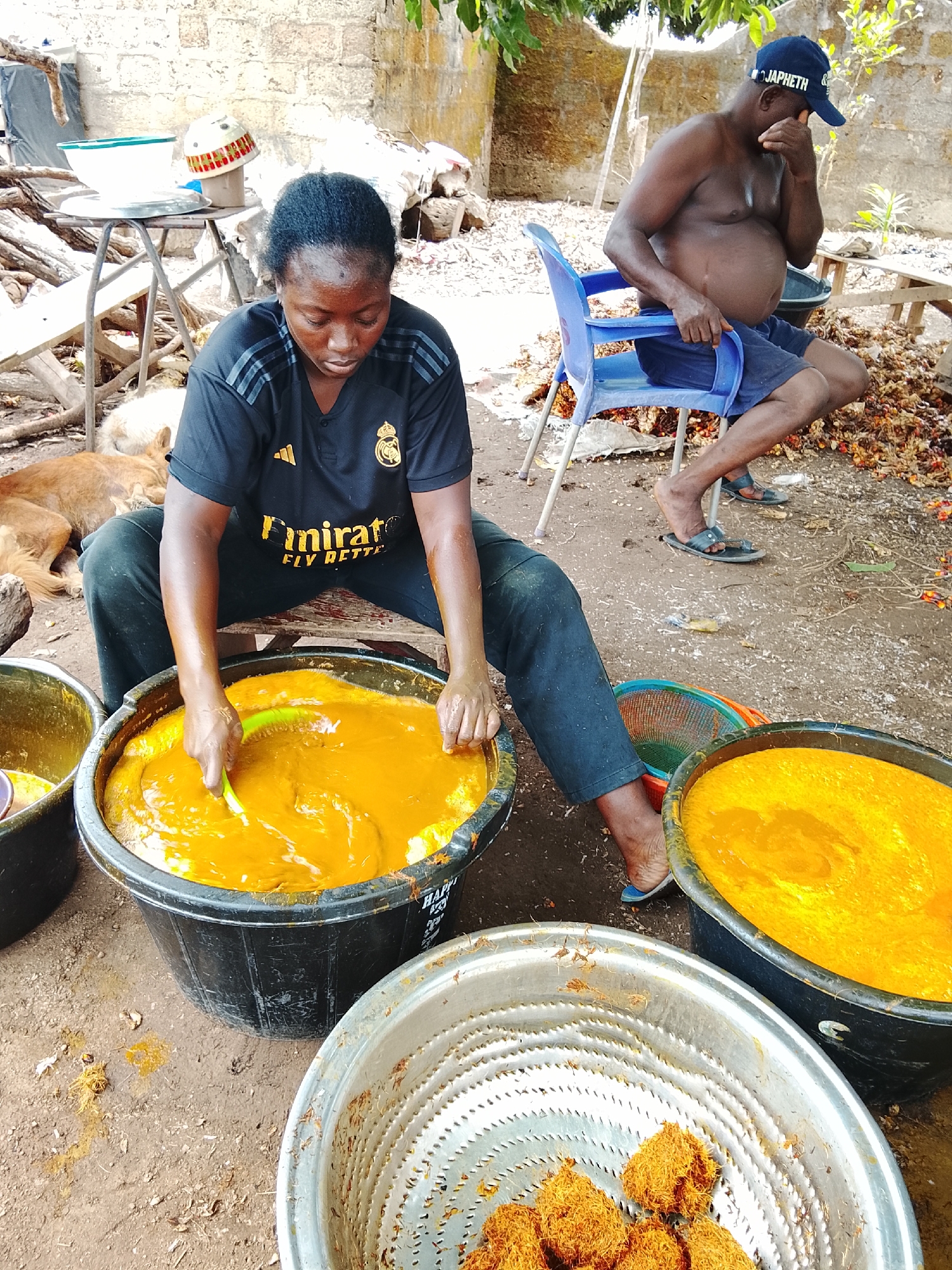 In Akwanga, Nigeria, a woman is seated, working with a large tub of bright orange liquid. She is wearing a black sports shirt and dark pants. Nearby, there's another similar tub and a metal bowl containing fibrous, orange-brown material. An older man sits shirtless on a blue plastic chair in the background wearing a cap and shorts. There's a dog sleeping near a table and some debris scattered on the ground.