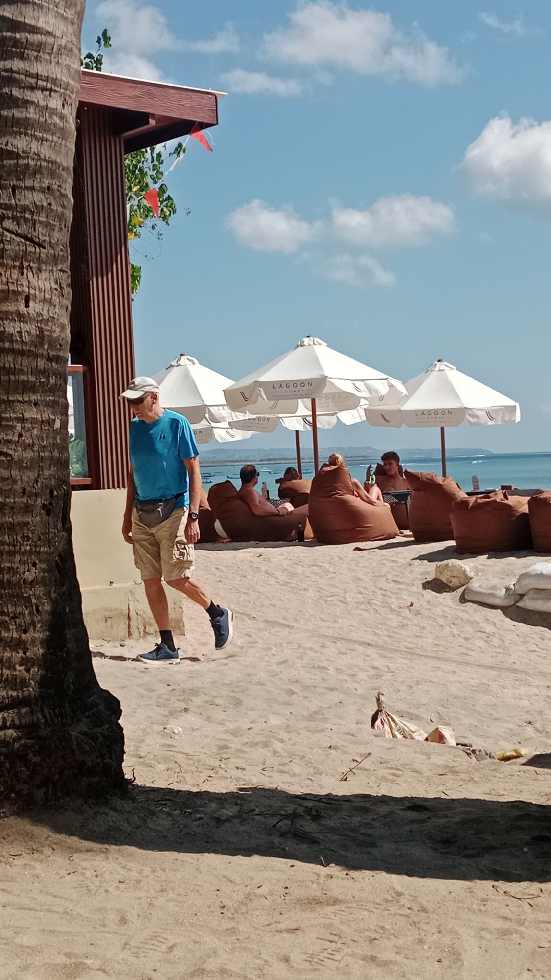 In Kuta, Indonesia, on a sandy beach, a man in a blue t-shirt, beige shorts, and a baseball cap walks from left to right. He is wearing a fanny pack around his waist and blue sneakers with white soles. In the background, several people are relaxing on large, brown beanbag chairs under white umbrellas. The umbrellas have "LAGOO LOUNGE" written on them. Behind the beachgoers, the ocean is visible with a distant coastline. A large palm tree trunk is in the foreground on the left side of the image. The sky is blue with scattered white clouds.