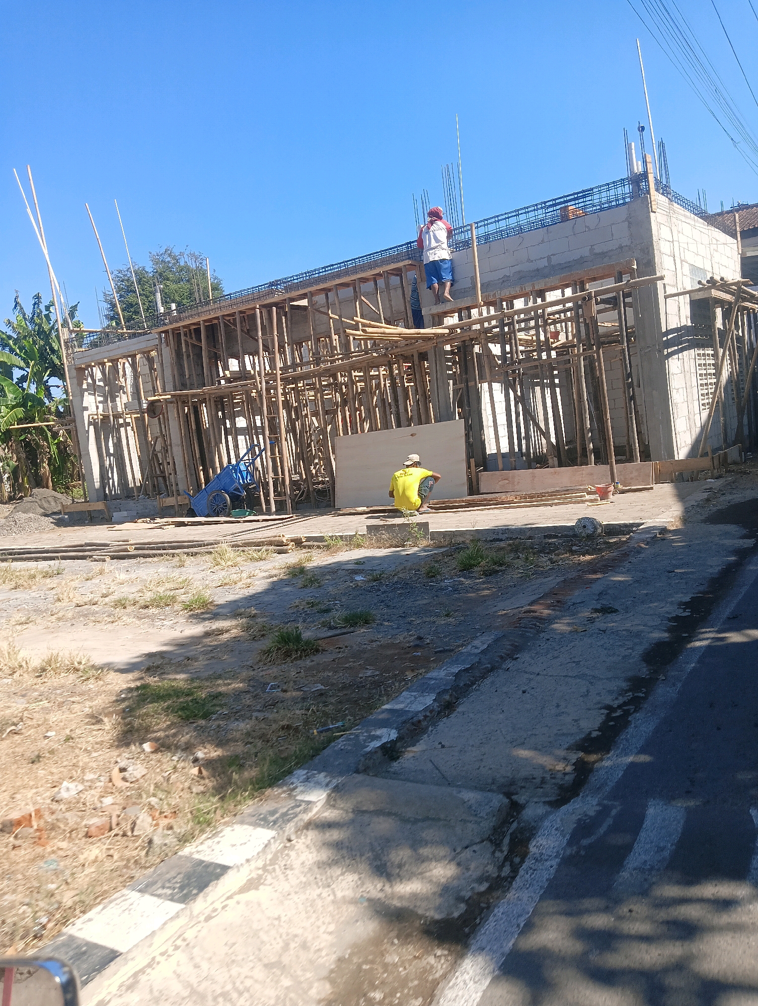 The image depicts a construction site in the city of Sokaraja, Indonesia, under a clear blue sky. A multi-story building is under construction, with wooden scaffolding supporting concrete and brick structures. Two construction workers are visible. One worker, wearing a red and white shirt and blue shorts, is standing on the second level of the building, near the edge. Another worker, dressed in a yellow shirt and dark pants, is seated on the ground level. A blue wheelbarrow is positioned near the seated worker. The surrounding environment shows dry vegetation, scattered construction debris, and a paved road in the foreground. Banana trees are visible in the background. The scene suggests ongoing construction work in an outdoor, possibly rural or suburban, setting within Sokaraja.