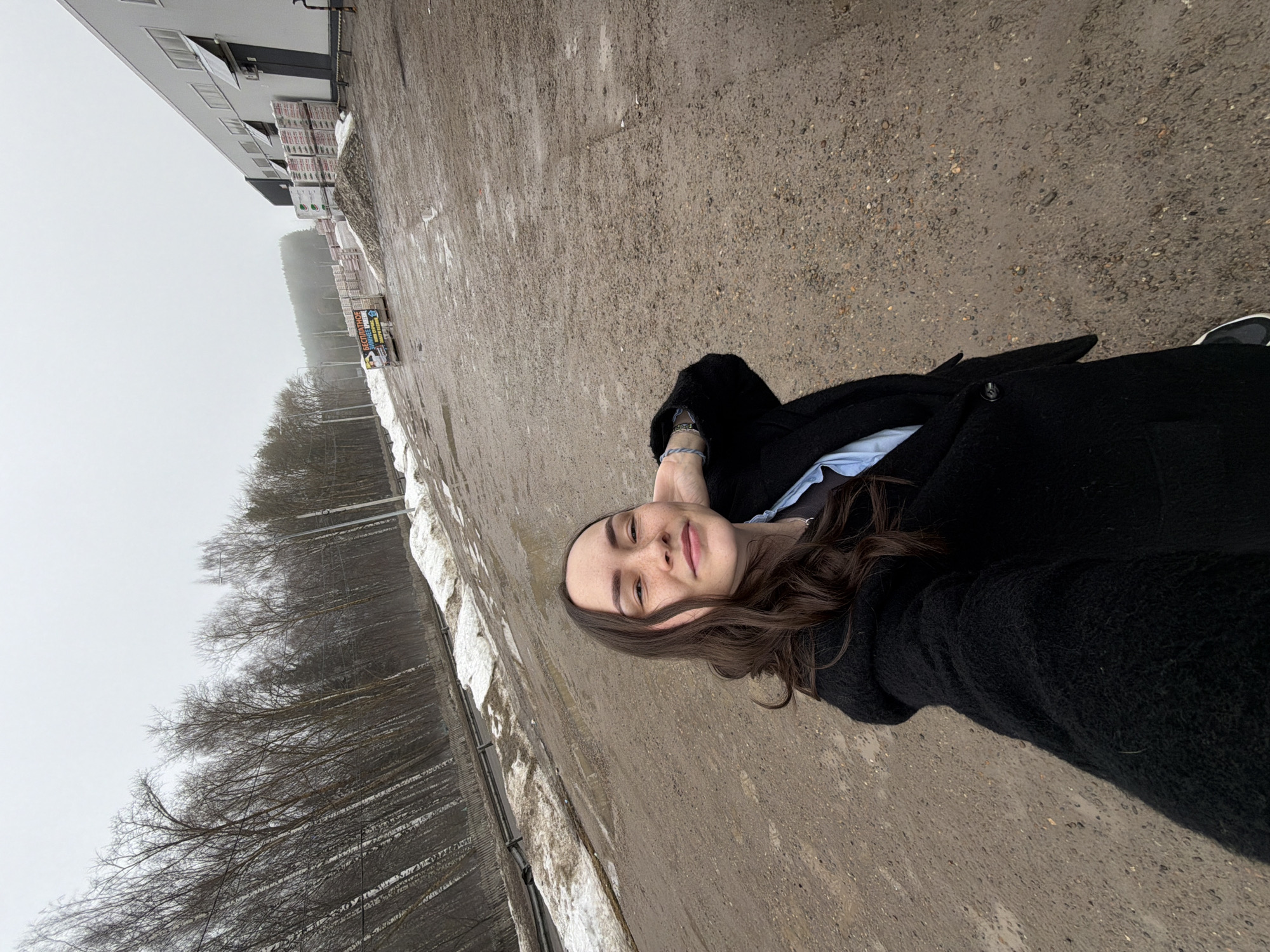 A woman lies on a concrete surface in Votkinsk, Russia. She wears a dark coat and has brown hair. Buildings and trees are visible in the background. The weather appears overcast. A small amount of snow is on the ground.