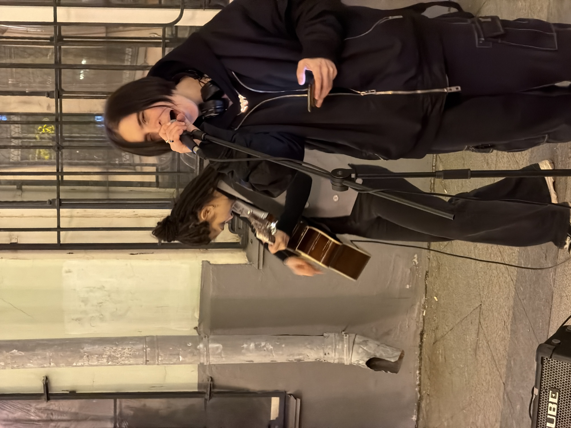 Two musicians are performing on the street, illuminated by streetlights. The foreground shows architectural details of the building behind them. The person on the upper part of the image sings into a microphone, wears a black hoodie and pants, and holds a smartphone. The lower person plays an acoustic guitar, wearing a black top. To the right, a "CUBE" amplifier is visible on the ground. The location is Moscow, Russia, as indicated by the architectural style and street elements.