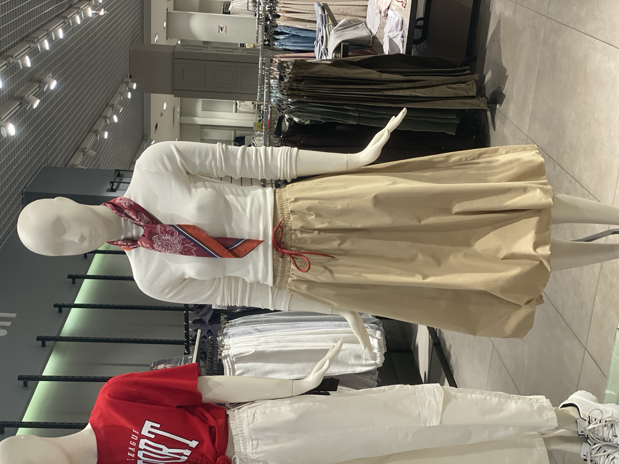 The image depicts two mannequins in a retail clothing store. The mannequin on the left is wearing a red t-shirt with white text and white cargo pants. The mannequin on the right is wearing a white long-sleeved shirt, a patterned scarf, and a beige skirt. Both mannequins are positioned in front of racks of clothing. The store environment includes tiled flooring, overhead lighting, and shelves displaying more apparel. The image was taken in the city of Novokuznetsk, Russia.