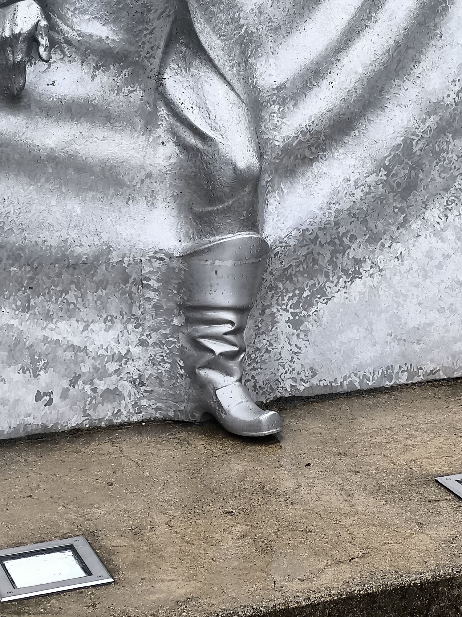 The image is a close-up, low-angle shot of the silver boot of a statue located in Kuldīga, Latvia. The boot is made of metal and appears to be part of a larger sculpture, likely depicting a historical or mythical figure due to the ornate detailing on the boot and the fabric-like texture of the statue's clothing. The boot is polished to a high sheen, reflecting light and creating highlights. The statue is positioned on a ground surface that appears to be concrete or stone, with visible aggregate. In the foreground, two square, recessed lights are embedded in the ground. The overall texture of the statue's clothing suggests a flowing or draped garment, adding to the grandeur of the piece. The lighting suggests it is daytime, with ambient light illuminating the scene.