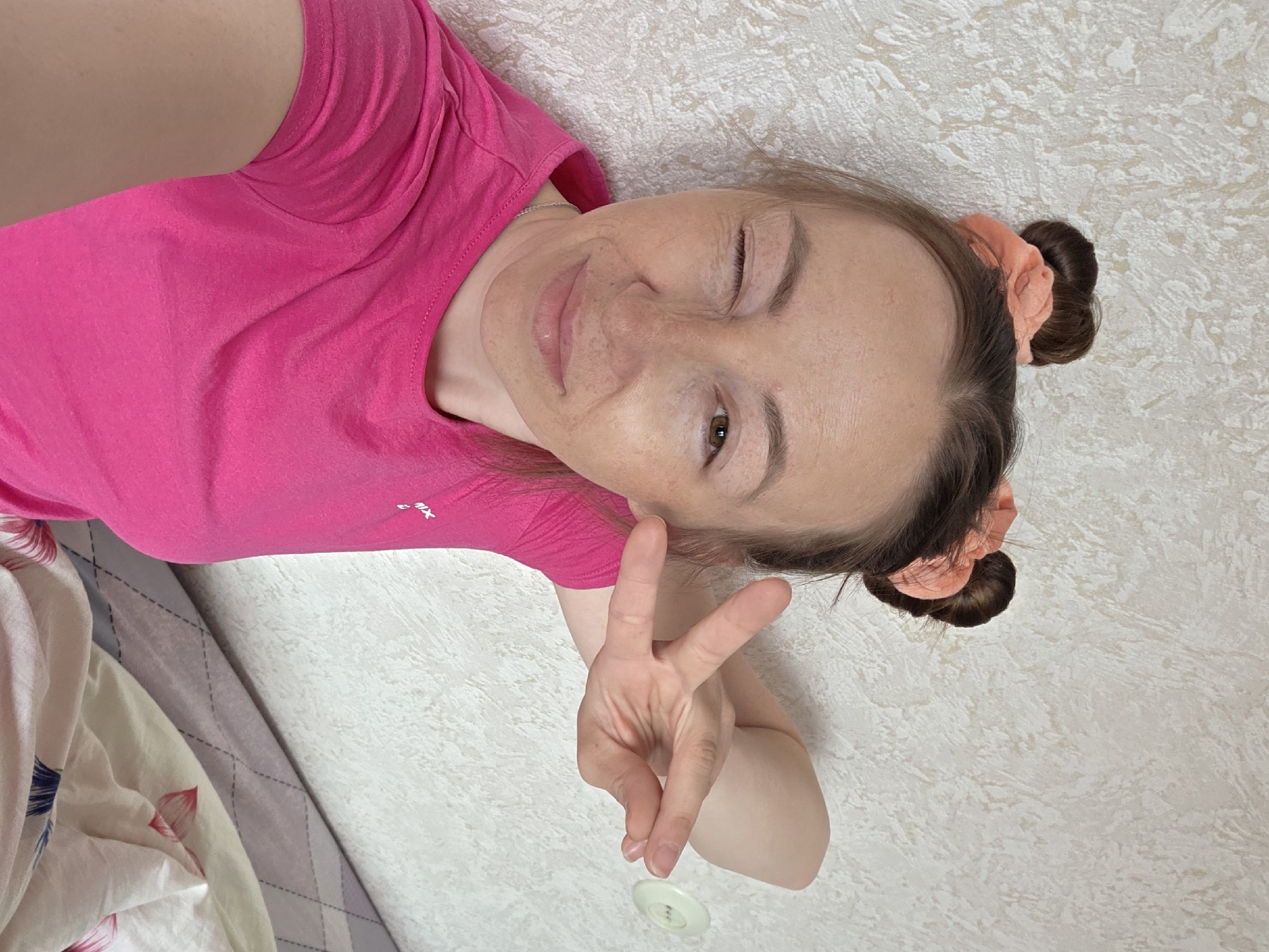 A woman is lying on a bed, winking at the camera and holding up a peace sign. She is wearing a pink t-shirt and has her hair in two buns with pink scrunchies. A white button lies near her hand. The bed has a white and gray patterned comforter. The wall is off-white with a textured pattern. This image was taken in Chaikovsky, Russia.