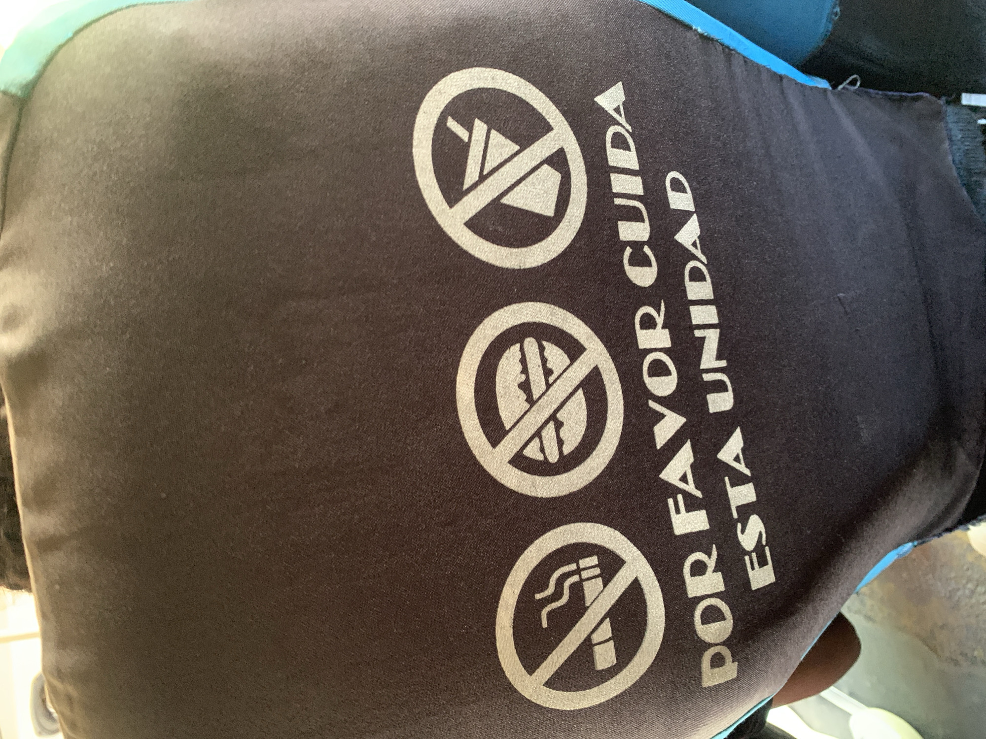The image captures a close-up of a dark gray or black fabric surface, likely the back of a seat or a protective covering within a public transport vehicle in La Asuncion, Venezuela. Three white circular "no" symbols are prominently displayed down the center of the fabric.
From top to bottom, the symbols depict:
1. A crossed-out cup or bottle with a straw, indicating "no drinks."
2. A crossed-out hamburger or sandwich, signifying "no food."
3. A crossed-out lit cigarette, representing "no smoking."
To the right of these symbols, white capital letters spell out "POR FAVOR CUIDA ESTA UNIDAD," which translates to "PLEASE TAKE CARE OF THIS UNIT." The text is oriented vertically along the right side of the frame.
A light blue or teal fabric trim is visible along the top and right edges of the dark fabric, suggesting it is part of the vehicle's upholstery or a seat cover. The fabric surface shows some minor texture and possible wear. The image is taken at an angle, focusing on these instructional signs, which convey rules for passengers within the vehicle. No specific activity is taking place, and no people are visible.