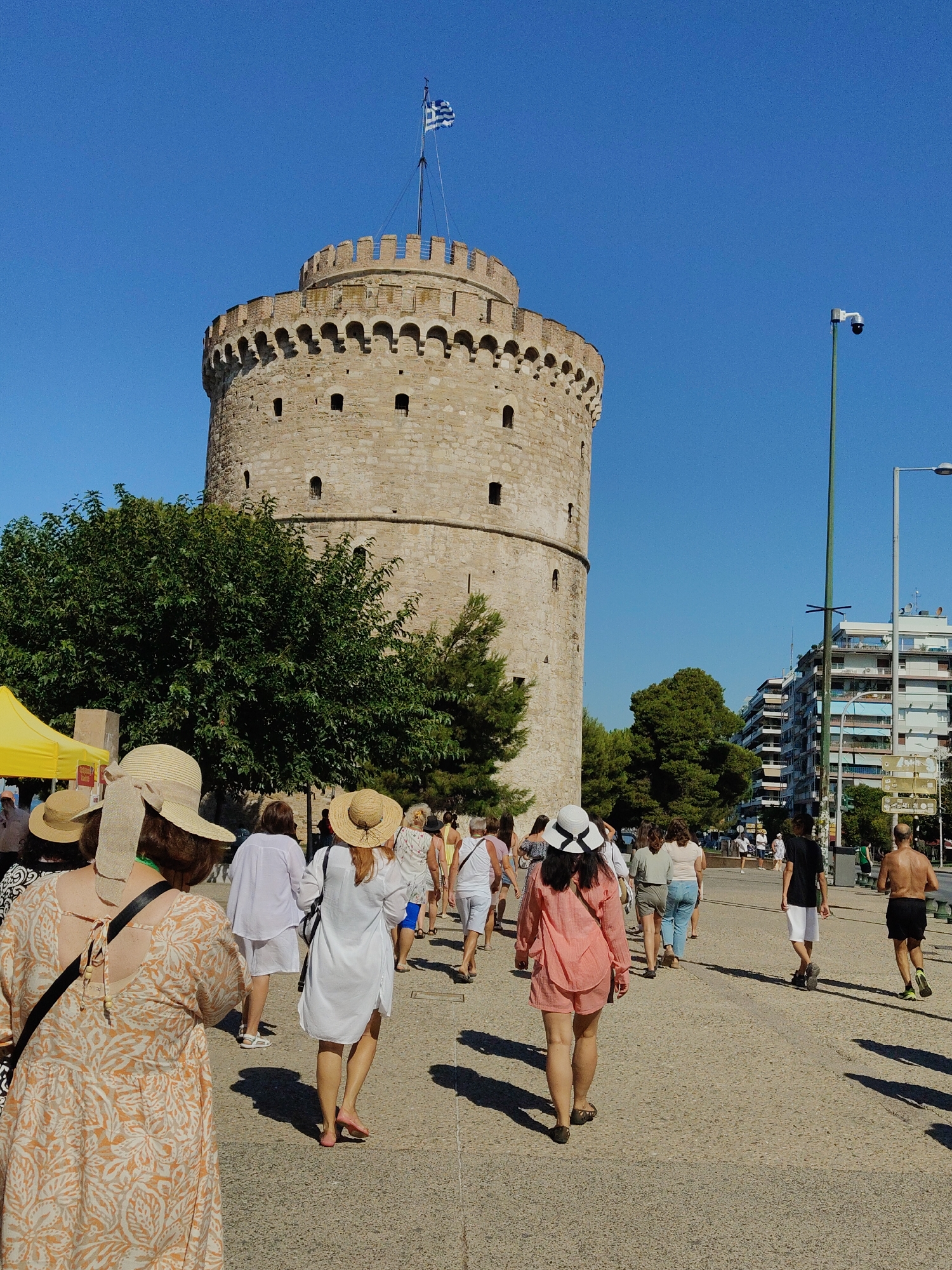 The image depicts the White Tower in Thessaloniki, Greece, a prominent landmark located in the Thessaloniki Municipal Unit. A group of people, mostly women wearing summer attire and hats, are walking away from the camera on a paved path. Behind them, the cylindrical stone tower stands tall against a clear blue sky, topped with the Greek flag. To the right of the tower, modern buildings and streetlights are visible, indicating an urban environment. The scene is bathed in sunlight, casting shadows on the ground. A yellow tent is partially visible on the left side of the image, suggesting a possible event or market. The overall atmosphere is that of a sunny day in a tourist area.