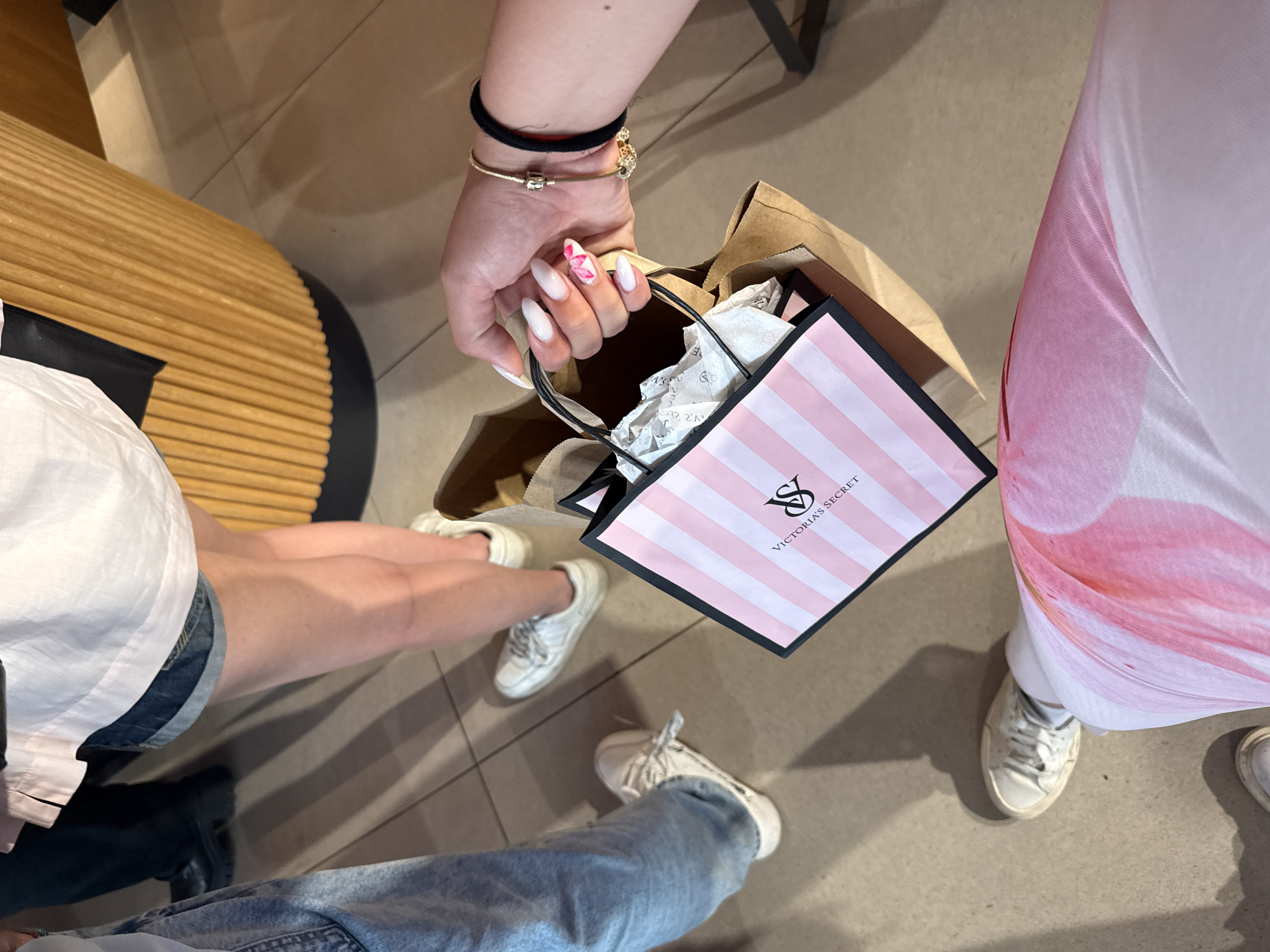 A high-angle shot captures a group of people standing indoors, likely within a retail environment such as a mall or store. The central focus is a hand holding a small Victoria's Secret shopping bag, characterized by its pink and white vertical stripes, black borders, and the "VS" logo with "VICTORIA'S SECRET" text. This striped bag is nestled within a larger, plain brown paper bag. The hand features long, white artificial nails, with a distinctive pink and white floral design on the ring finger. Gold bracelets and a black hair tie are visible on the wrist.
Around the central image, the lower bodies and legs of at least three other individuals are partially visible. To the left, a person wears denim shorts, a white top, and white sneakers, standing near a curved wooden slatted bench or decorative element. To the right, another individual is seen wearing a flowing garment with a pink and white pattern, paired with white sneakers. At the very bottom of the frame, a third person's lower legs are visible, clad in blue jeans and white sneakers.
The setting features a light-colored, possibly tiled, floor with visible grout lines. Distinct shadows are cast on the floor, suggesting indoor artificial lighting. The scene conveys a casual shopping atmosphere.