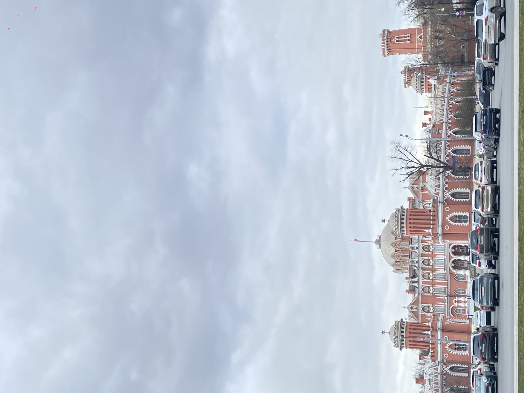 Here is a description of the image:
The image shows a row of cars parked next to a distinctive, ornate red-and-white building. The building features a dome and several towers. The sky is overcast. The image is taken along a city street in Khimki, Russia.