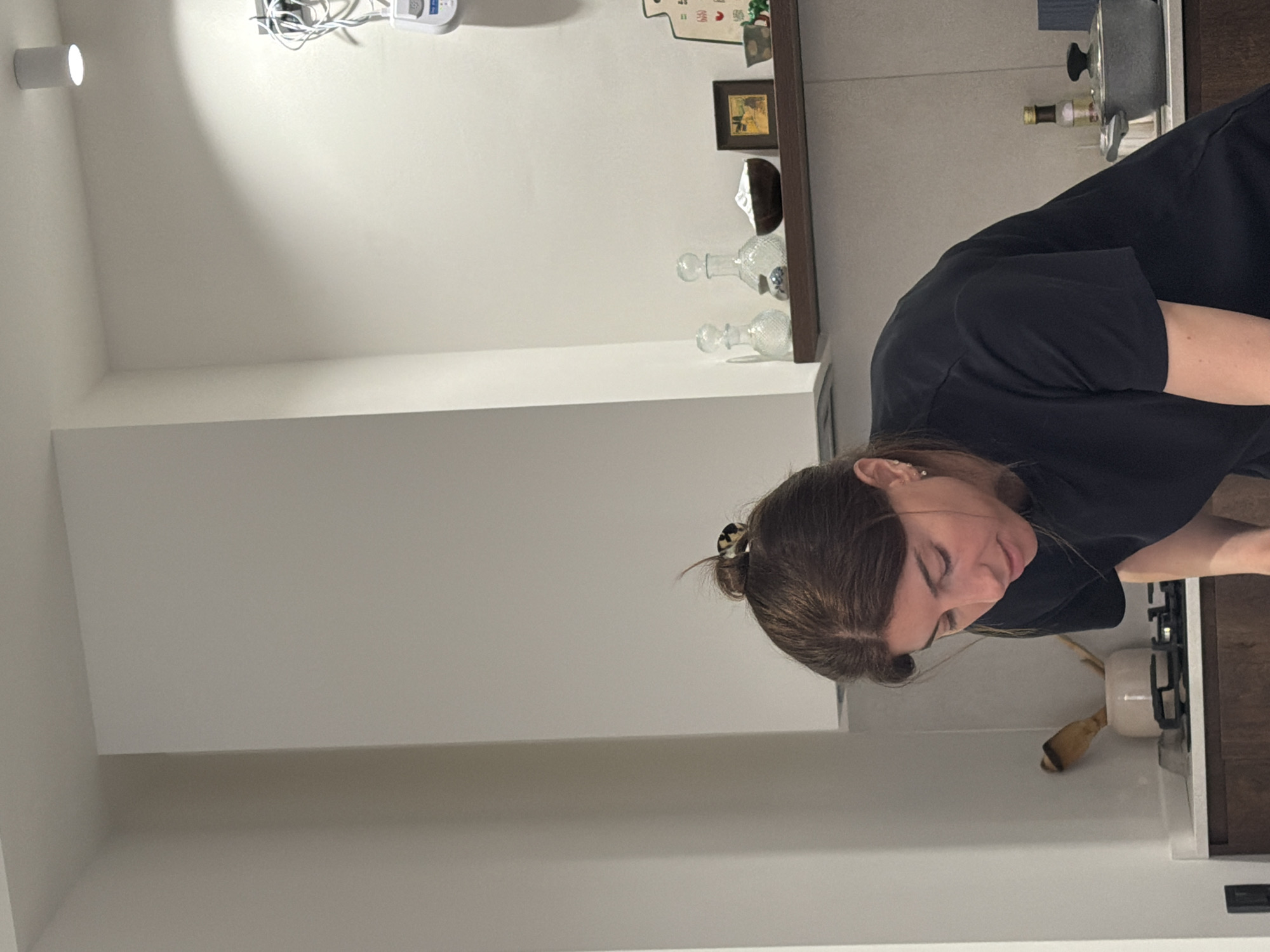 A woman in a black t-shirt is standing in a kitchen. She has brown hair pulled back in a bun. Behind her are a stove, pots, cooking utensils, and a shelf with decorative bottles. The location is potentially a residential kitchen, but the specific location in Tbilisi, Georgia, is not discernible from the image.