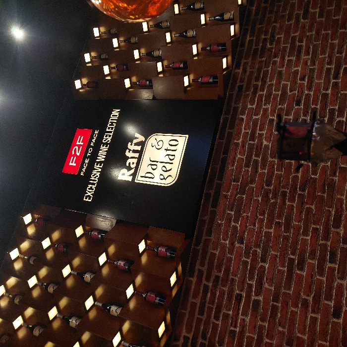 This image depicts the interior of a Raffy Bar & Gelato establishment in the city of Plovdiv, Bulgaria. A large, illuminated black sign is centrally positioned in the upper half of the frame. The sign prominently features "F2F" in a red box with "FACE TO FACE" written below it, followed by "EXCLUSIVE WINE SELECTION." Below this, "Raffy" is displayed in a script font, and "bar & gelato" is enclosed within an illuminated oval.
Flanking the central sign are dark wooden shelving units configured in a grid pattern. Each compartment of these units holds a single wine bottle and is individually illuminated by a warm, recessed light from below. Approximately 25-30 wine bottles, varying in label and bottle color, are visible across these displays.
The lower half of the scene is dominated by a reddish-brown brick wall with light-colored mortar, laid in a stretcher bond pattern. In the lower right corner, a dark object with a metallic top, covered in foil, is partially visible, consistent with the appearance of a hookah. The overall lighting is artificial, with targeted illumination on the signage and wine displays, supplemented by overhead lighting. No individuals are present in the image.