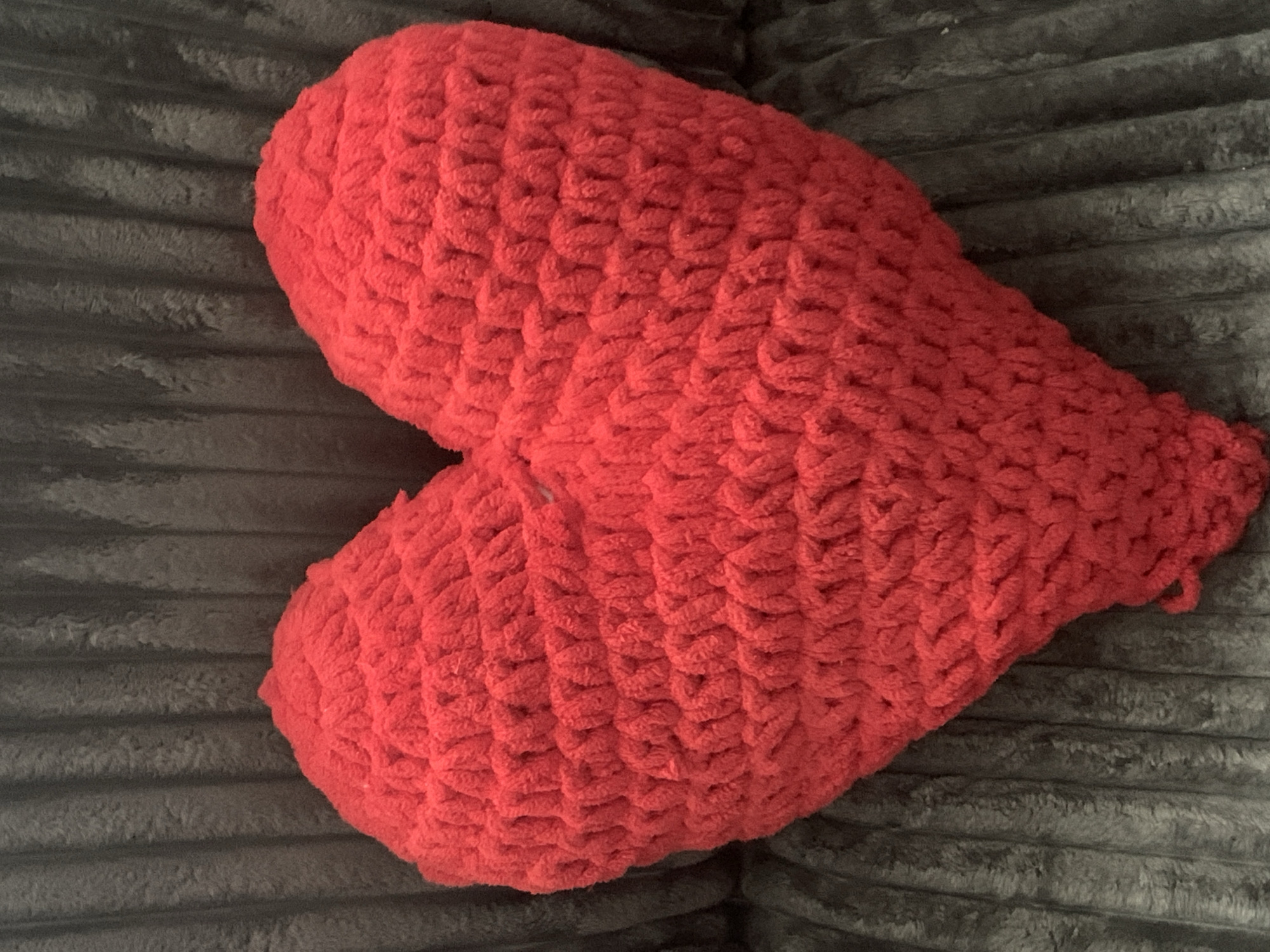 The image shows a red, heart-shaped crocheted pillow placed on a dark gray, textured surface, likely a sofa or blanket. The pillow is the central focus, filling most of the frame. The texture of the crochet stitches is clearly visible, creating a soft, plush appearance. The background surface has a ribbed or furrowed texture, adding a contrasting visual element to the smooth curves of the pillow. There are no people or other objects present in the image. There is no information within the image that would allow for identification of its location in Tashkent, Uzbekistan.