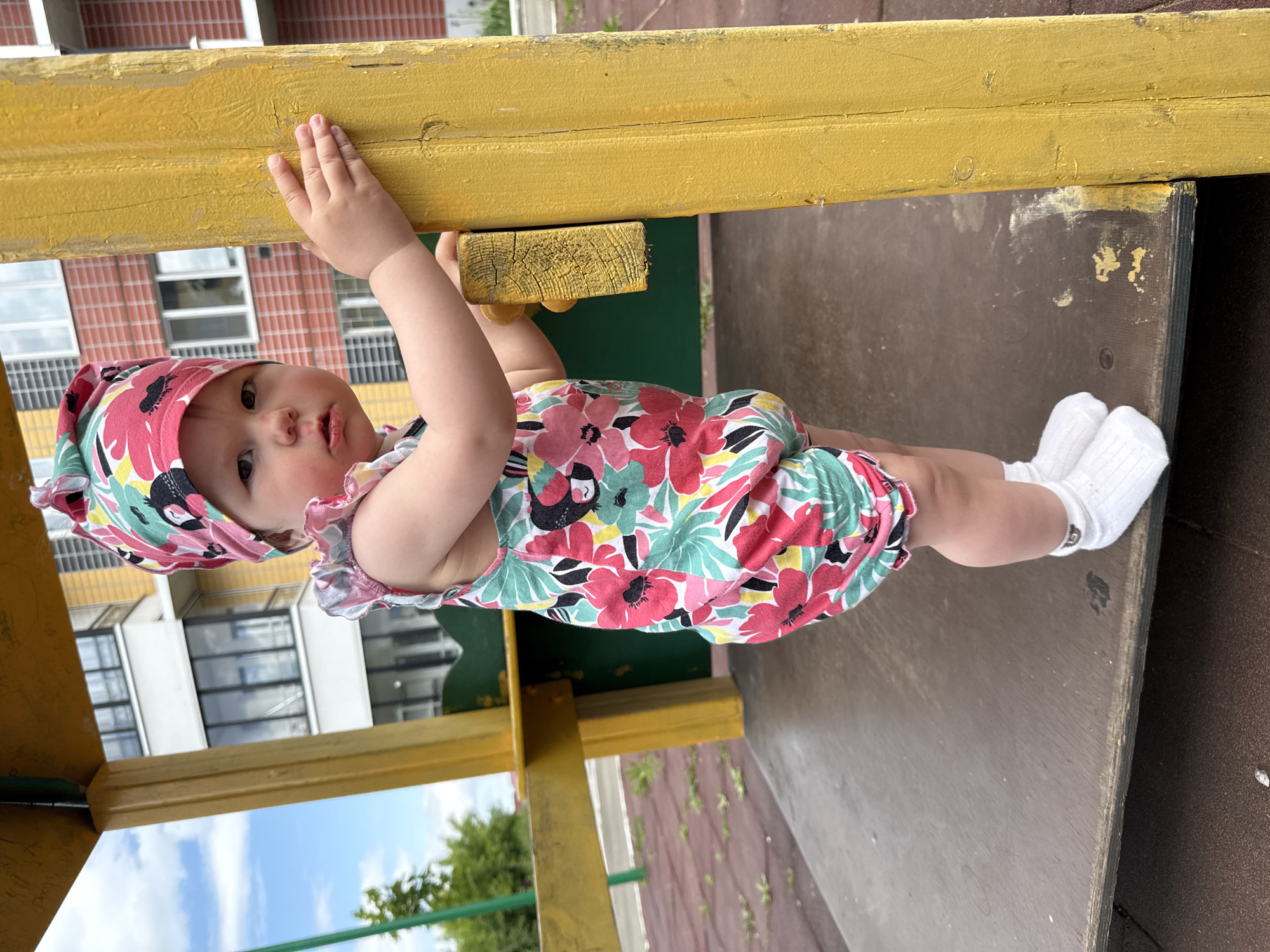 A young Caucasian child, appearing to be around one year old, stands on a wooden platform of a playground structure. The child is wearing a colorful floral print romper and a matching bandana with a bird motif. They have white socks on their feet. The child is facing forward, looking towards the camera, with their right hand resting on a weathered yellow wooden support post. Behind the child, a modern apartment building with a red and yellow facade is visible, suggesting an urban or suburban setting. The playground surface appears to be a dark material, possibly rubber or asphalt. The sky is cloudy. The overall impression is of a candid outdoor photograph of a child on a playground.