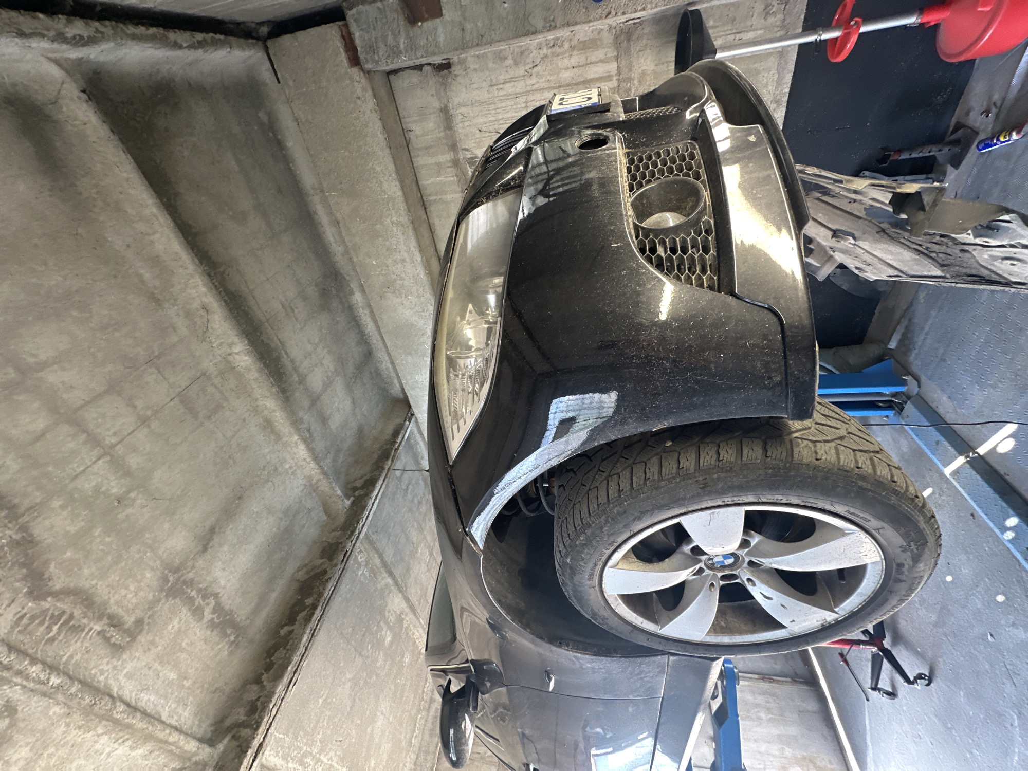 The image shows the front-left side of a black BMW car, possibly undergoing maintenance or repair. The car is elevated, suggesting it is on a lift or jack, revealing its underside and front wheel. The wheel is a silver BMW alloy with a winter tire. The car's headlights and grille are visible. The setting appears to be a garage or workshop, with concrete walls and ceiling. There are various tools and equipment in the background, including a red oil drain pan, a red and silver tool, and blue hydraulic lift arms. The lighting suggests it is daytime. The image is taken from a low angle, looking upwards at the car. No people are visible. The visible text on the license plate frame reads "EU".