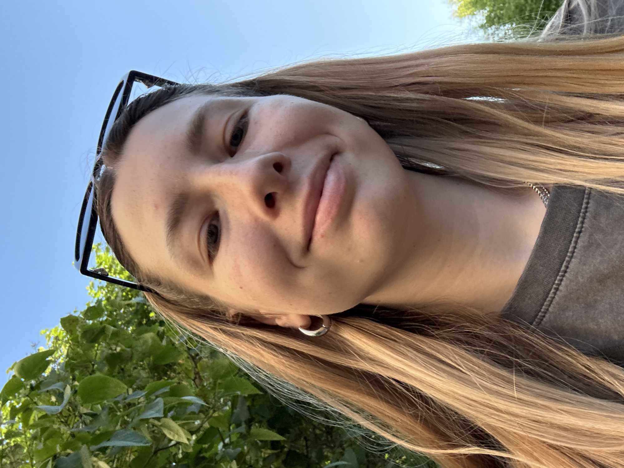 A young adult female is depicted in a close-up, upward-angled selfie, set outdoors in the city of Moscow, Russia. Her long, straight, light brown hair with prominent blonde highlights is parted down the middle and cascades over her shoulders. She wears black-framed sunglasses pushed atop her head, resting on her hair. Her face, viewed from below, features a subtle, closed-mouth smile with naturally colored lips, a small nose, and brown eyes looking slightly downward towards the camera. Her skin appears fair with faint freckles visible on her nose and cheeks. A single, thick silver-toned hoop earring is worn in her left ear (viewer's right). A thin, silver-colored ball chain necklace is partially visible on her right side, beneath her hair. She is dressed in a dark gray, possibly acid-washed or faded, crew-neck t-shirt with visible stitching along the collar. The background on the left features dense, vibrant green foliage from trees, while the right side of the frame is dominated by a clear, bright blue sky, indicating sunny daytime conditions.