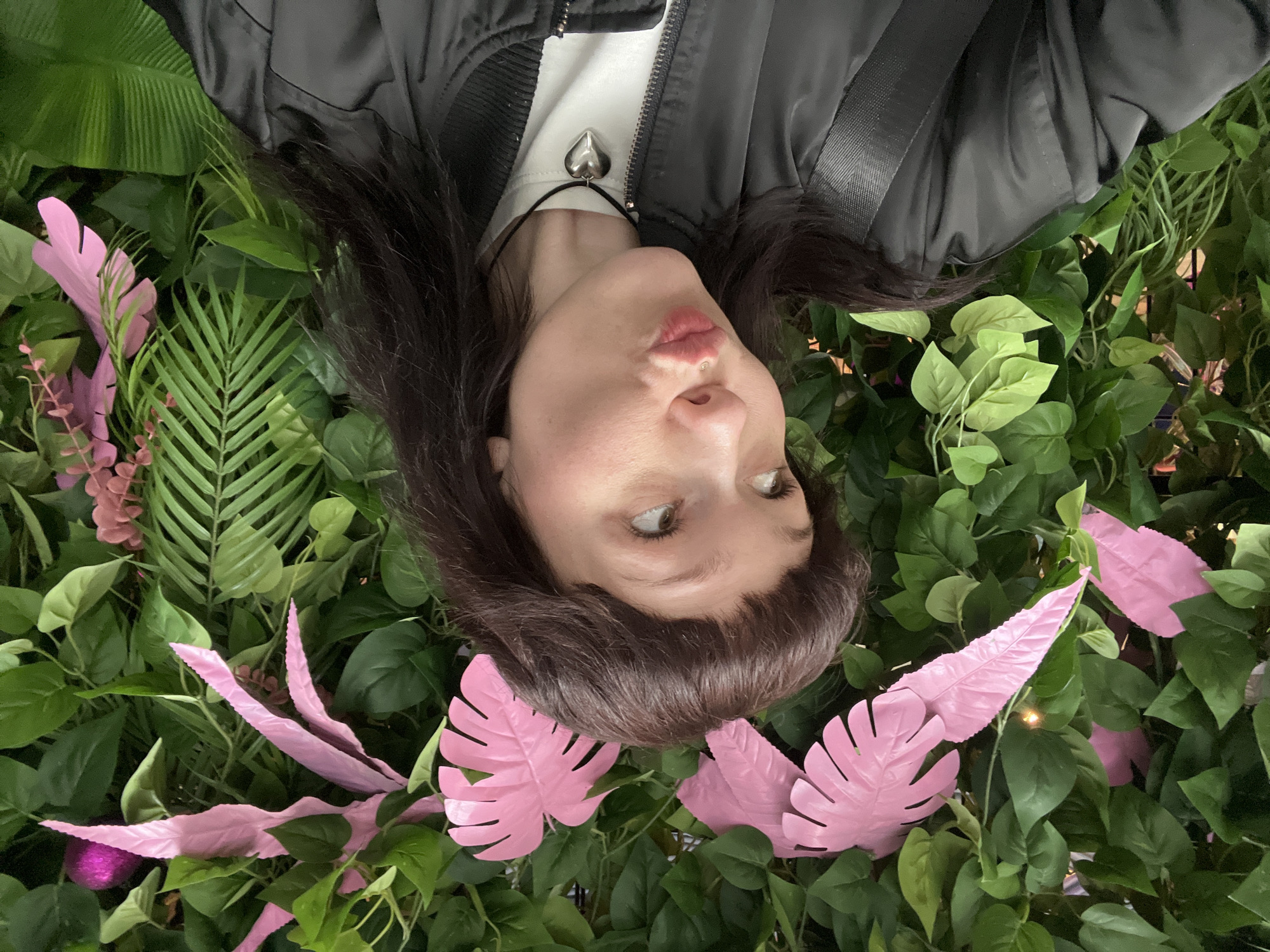 A woman is lying on her back among artificial foliage that includes green leaves and pink monstera-like leaves. She has brown hair with bangs and is wearing a black bomber jacket over a white shirt with a silver heart-shaped pendant necklace. She is looking slightly to her left with pursed lips. The scene may be an artificial display or photo opportunity. The location is unknown and cannot be confirmed from the image.
