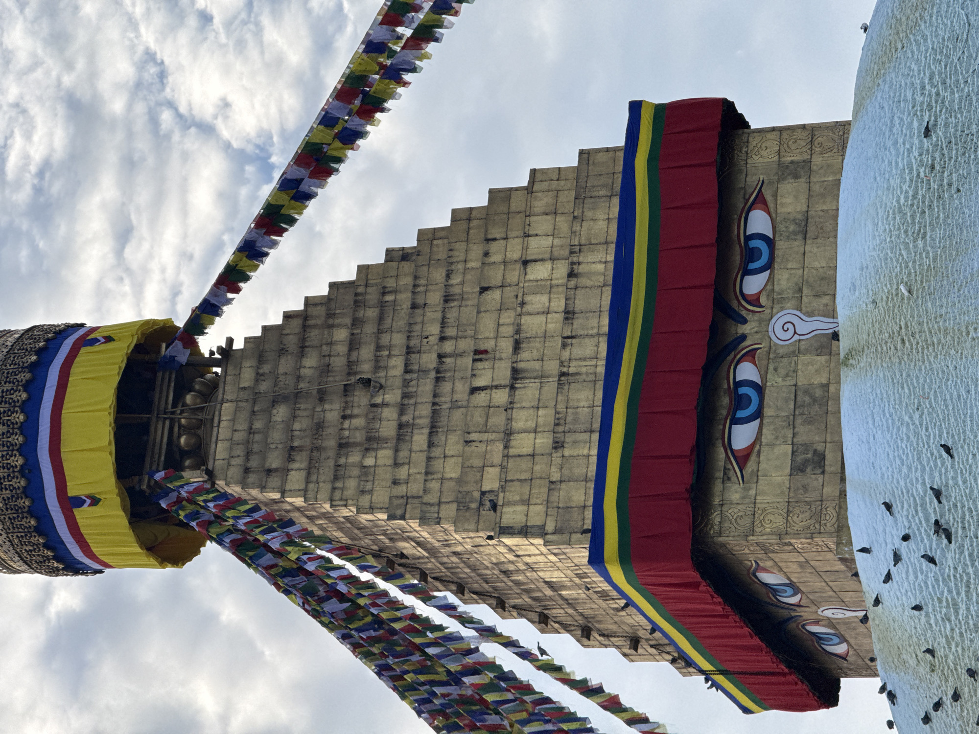 The image shows the Boudhanath Stupa in Kathmandu, Nepal, under a cloudy sky. The stupa, a prominent landmark, is adorned with prayer flags strung across its upper sections. Large, painted eyes, a common feature of stupas in Nepal, are visible on the structure. The lower portion of the stupa appears to be covered in a light dusting of snow or frost, with several pigeons scattered across this surface and the surrounding area. The stupa's golden facade is prominent, and bands of red, yellow, and blue fabric are wrapped around its upper levels.