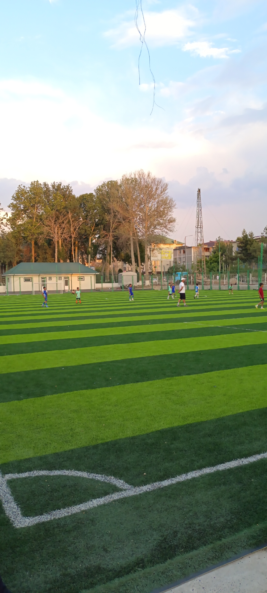 An outdoor artificial turf soccer field is depicted at dusk in Fergana City, Uzbekistan. The field is striped with alternating dark green and light green turf. Several individuals, likely children and at least one adult coach, are engaged in a soccer practice. Some individuals are in motion, suggesting active participation in drills or a game. In the background, trees line the perimeter of the field, behind which are buildings and structures. The sky is partly cloudy with a blend of blue and soft pink hues, indicative of the approaching evening. A thin, dark, string-like object hangs suspended in the upper portion of the frame against the sky.