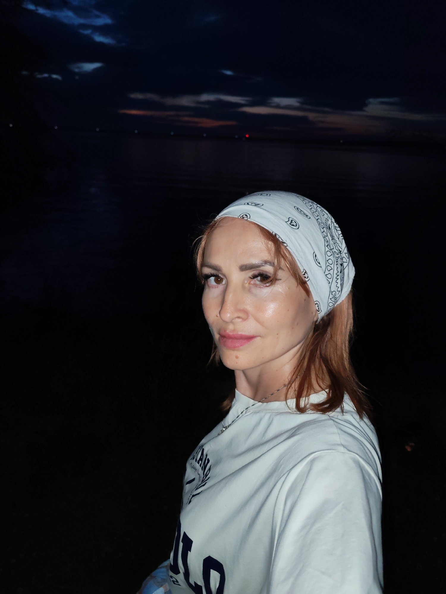 A medium shot shows a woman with red hair wearing a white bandana with a black paisley pattern and a white t-shirt with text. She is standing outdoors at dusk, with a dark body of water and a dimly lit sky in the background. The water appears to be a lake or river, reflecting the fading light. The overall scene is dark and atmospheric. The location is identified as the city of Chaikovsky, Russia.