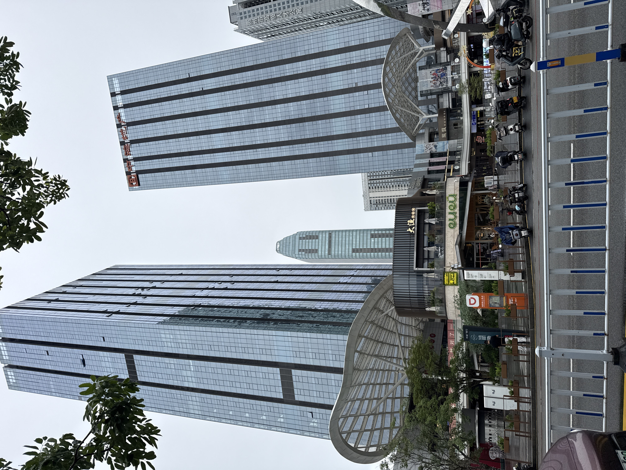 The image captures a street-level view of a modern urban commercial district in Huizhou, China. Two prominent glass-clad skyscrapers dominate the background, with one featuring orange lettering on its upper facade. In the foreground, a multi-level shopping complex with a distinctive curved, translucent roof structure is visible. Several parked motorcycles and scooters line the street in front of the complex, some with riders. People are seen walking on the sidewalk and near the entrance of the shops. Signage in Chinese characters is present on the buildings and on various street furniture, including what appears to be a digital information kiosk and directional signs. The sky is overcast, suggesting a cloudy day. The scene depicts a bustling commercial area with a mix of large architectural structures and everyday street activity.