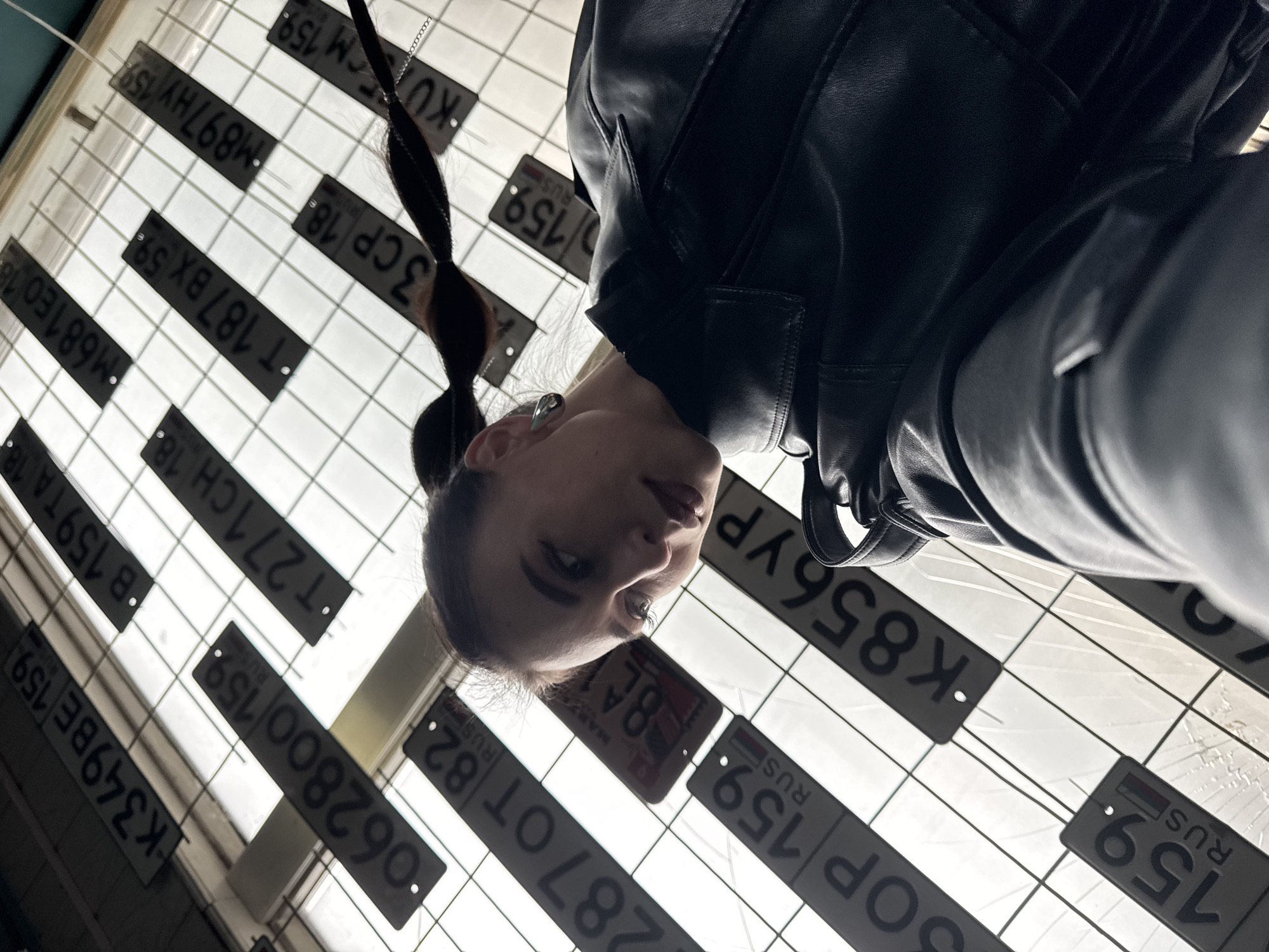 The image presents a unique low-angle portrait of a young woman set against a striking background of suspended Russian license plates. The woman, with dark hair styled in a ponytail, wears a black leather jacket. Her head is positioned in the lower part of the frame, and she gazes directly at the camera with a neutral, slightly pensive expression.
The background features a grid-like structure, likely a translucent ceiling or panel, from which numerous dark gray license plates are hung. Bright light emanates from behind these plates, casting them in silhouette and creating a dramatic contrast with the woman's dark clothing. This arrangement gives the scene an urban, modern, or possibly industrial aesthetic, suggesting an indoor display or installation.
Numerous Russian license plate numbers are clearly visible, all bearing the "RUS" country identifier and various regional codes. Examples include "K349BE 159 RUS", "062800 159 RUS", "287 OT 82 RUS", "SOP 159 RUS", "K856YP RUS", "B 159 TA 18 RUS", "T271CH 18 RUS", "M681E0 18 RUS", "T 187 BX 59 RUS", "3CP 18 RUS", and "M897 HY 159 RUS". The frequent appearance of region codes like "159" (Perm Krai) and "18" (Udmurt Republic) aligns with the location context of Chaikovsky, Russia. The upward perspective and the unique backdrop create an engaging and artistic composition.
