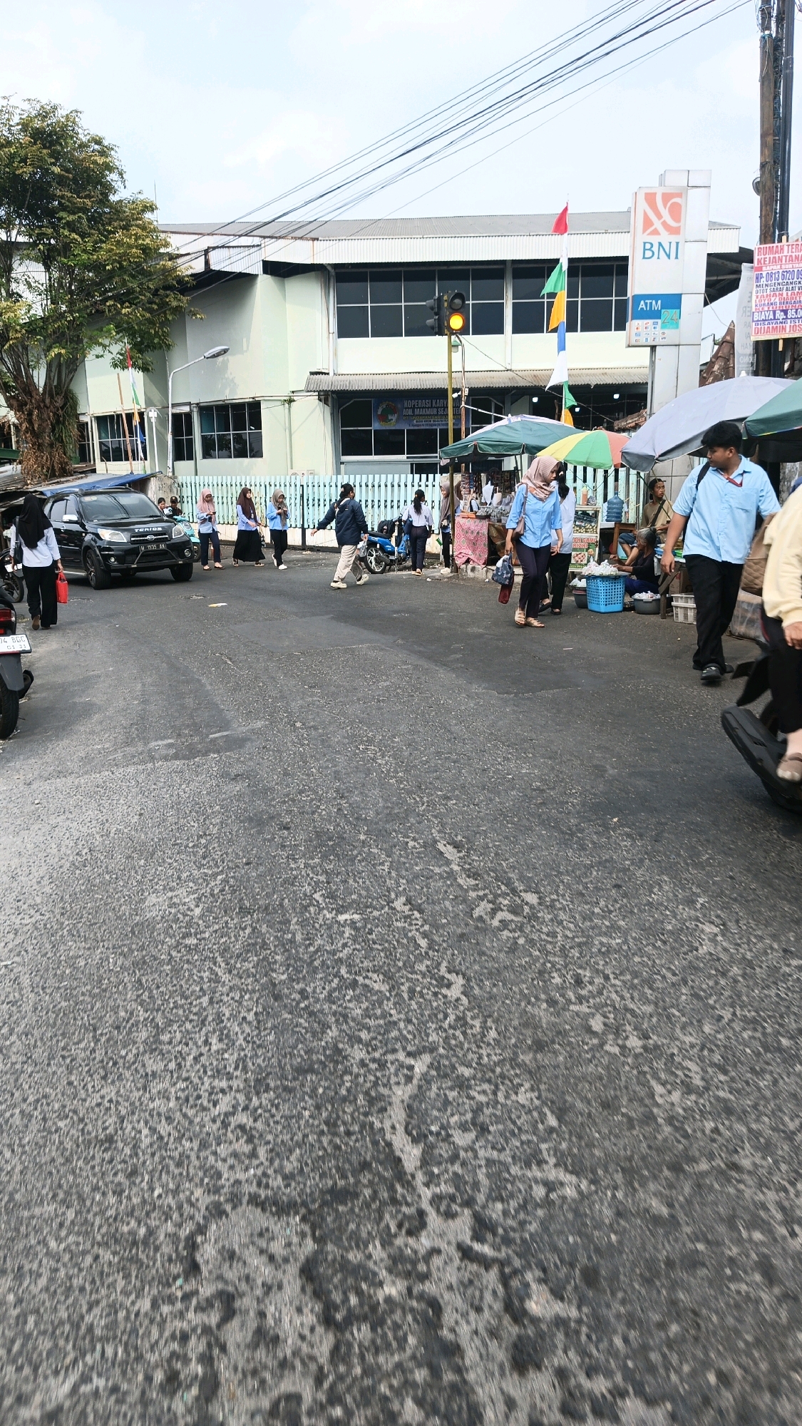 A street scene unfolds in Genuk Barat, Indonesia, characterized by a light-colored building with large windows and a BNI ATM located on its facade. A traffic light, currently displaying amber, signals a potential change in traffic flow. Several individuals are present, some of whom appear to be engaging in commercial activities under market stalls adorned with colorful umbrellas. A black SUV is parked on the side of the road, and a motorcycle is visible as well, suggesting a bustling urban environment. The road surface is asphalt, showing signs of wear and tear. The presence of multiple people, vehicles, and commercial activity indicates a lively public space.