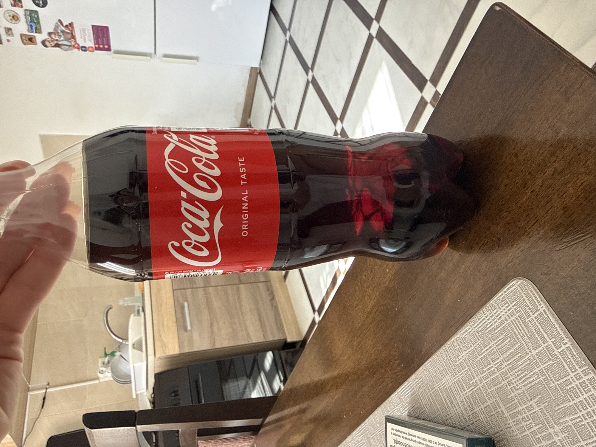 A Coca-Cola bottle is held by a person's hand above a wooden table. The bottle is full of dark liquid. In the background, a kitchen counter with a sink and cabinets is visible, along with a white refrigerator with magnets on it. The floor is tiled with a checkerboard pattern. The scene appears to be indoors, likely a home kitchen. The overall lighting is bright, with sunlight casting shadows on the floor.