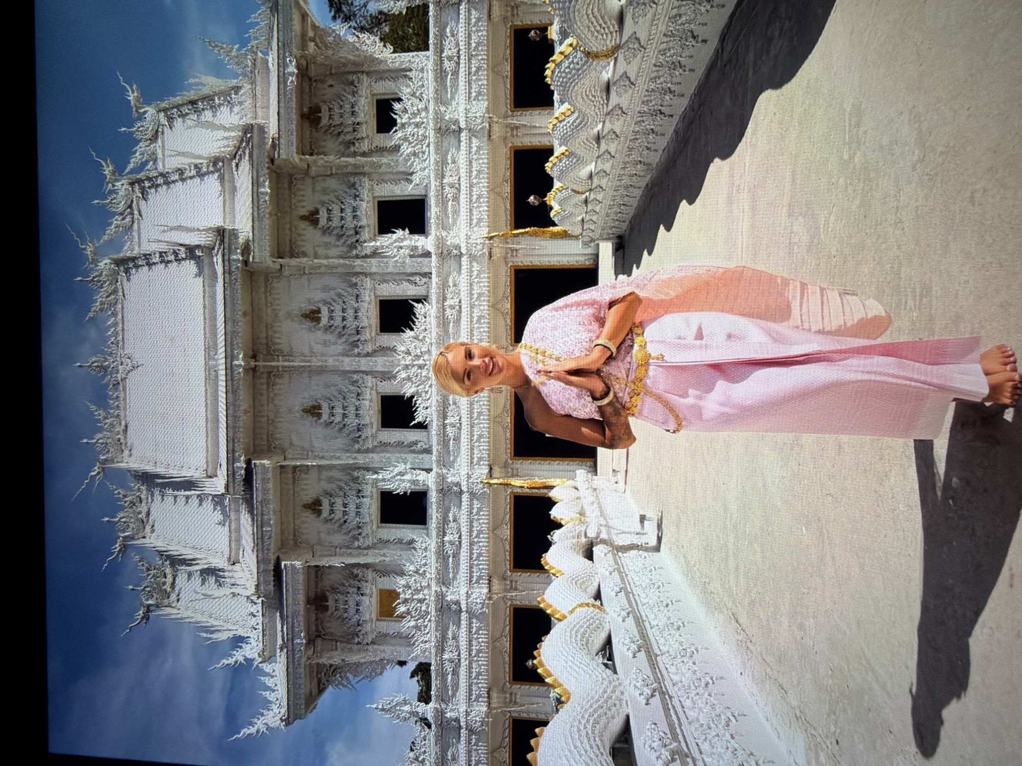 An individual with blonde hair and a radiant smile is captured in a full-length portrait, performing a traditional *wai* gesture with hands pressed together at chest level. They are adorned in a traditional Thai ensemble, predominantly pink with intricate gold detailing, including a multi-layered skirt (pha nung or pha sin) and a fitted top, complemented by golden jewelry. The person is barefoot.
The setting is outdoors, under a clear blue sky with bright sunlight, which casts distinct shadows on the light-colored ground. Behind the individual stands an exquisitely ornate white temple structure, strongly resembling Wat Rong Khun (the White Temple) in Thailand. The temple's architecture is highly detailed with multiple tiers, elaborate sculptural elements, and Naga-like balustrades finished with gold accents. The intricate carvings and spires are prominent against the blue sky.
The scene depicts a pleasant moment, likely a tourist or visitor posing at a significant cultural landmark. While the image's "location context" is noted as Hamburg, Germany, the visual elements unequivocally point to a location in Thailand, specifically its distinctive temple architecture and traditional attire. There is no visible text in the image.