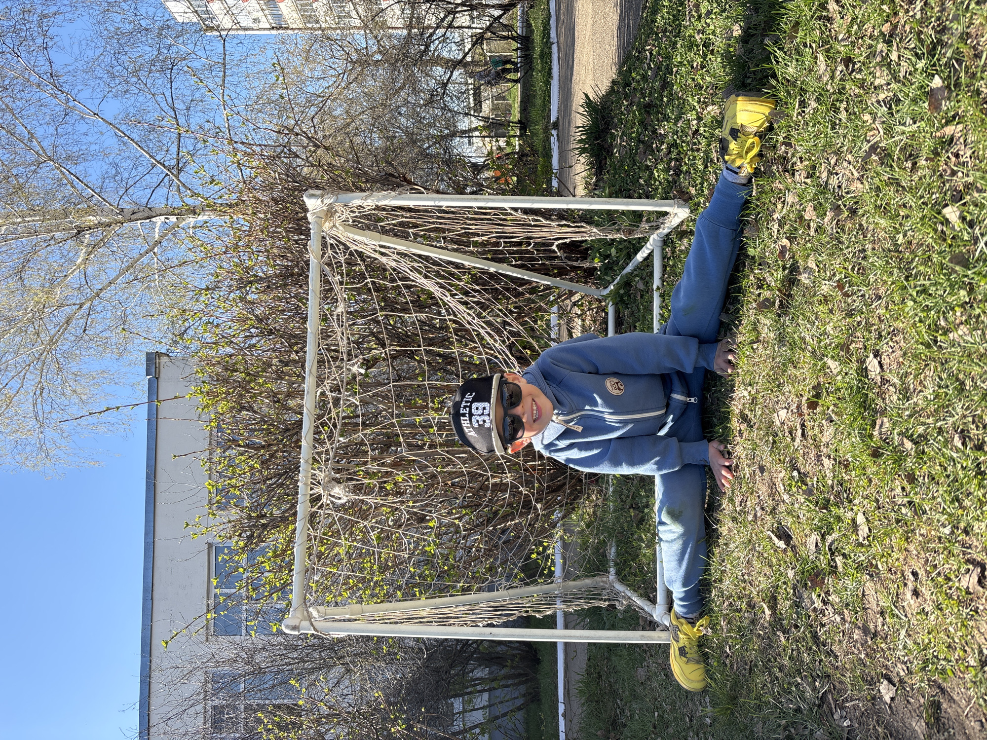 A child wearing a blue sweatsuit, a black hat with "ATHLETIC 33" printed on it, and yellow sneakers is positioned in front of a small white soccer goal with a disheveled net. The child is smiling at the viewer. They are on a grassy area with patches of dirt and dead leaves. Behind the goal, there are bushes and trees, and in the background, there are buildings. The sky is blue. The location is in the city of Chaikovsky, Russia.