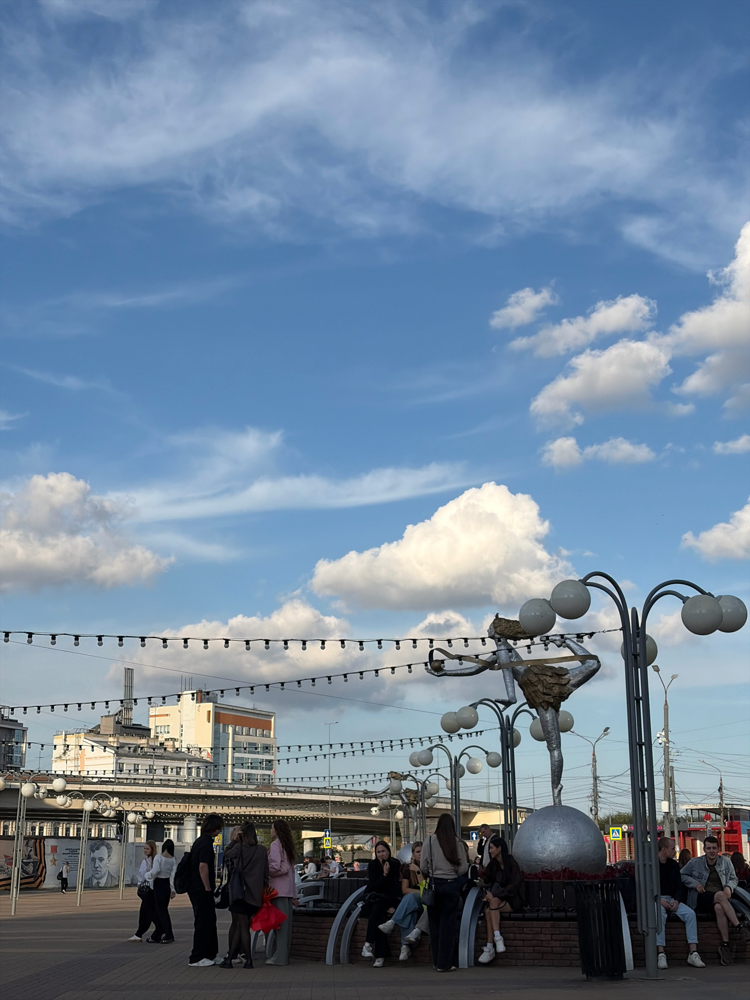 This photograph captures a street scene in Nizhny Novgorod, Russia. In the foreground, a group of young people are gathered, some sitting on a brick structure, others standing. Among them, a person wearing a pink jacket and holding a red bag is visible. To the right, a group of young men are also seated, with one wearing a denim jacket. In the background, a statue of a figure balancing on a sphere is prominent, surrounded by decorative streetlights with spherical globes. Further in the background, buildings and an overpass are visible, with a string of lights stretching across the frame above the people. The sky is filled with a mix of scattered clouds and wispy cirrus formations against a blue backdrop. The ground is paved with cobblestones or similar material. The overall atmosphere suggests a public gathering space in an urban environment.