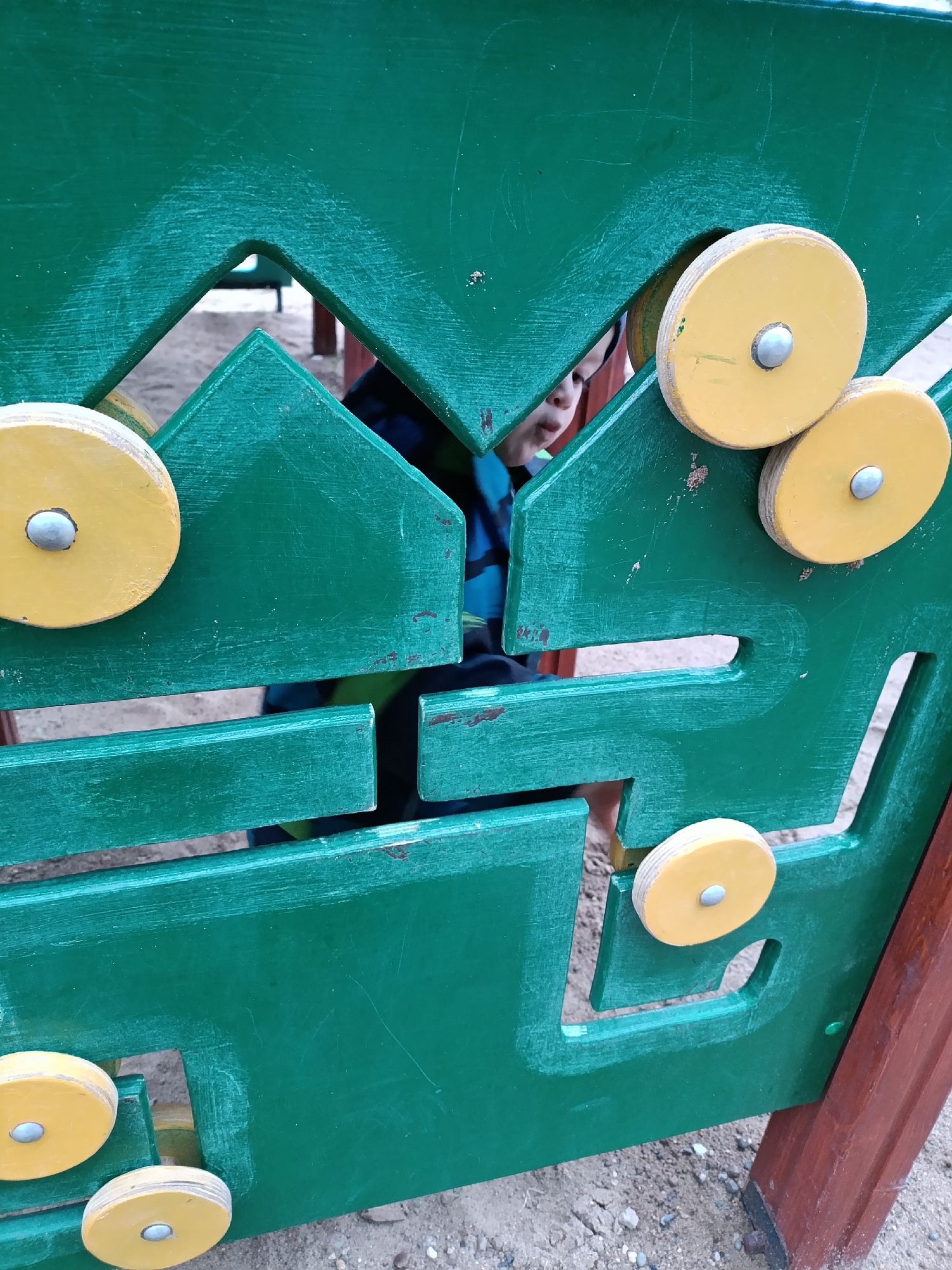 A child peeks through a green wooden play structure in Chaikovsky, Russia. The structure features geometric cutouts and yellow circular knobs with metal fasteners. The ground is sandy. The child is wearing a blue and black striped shirt.