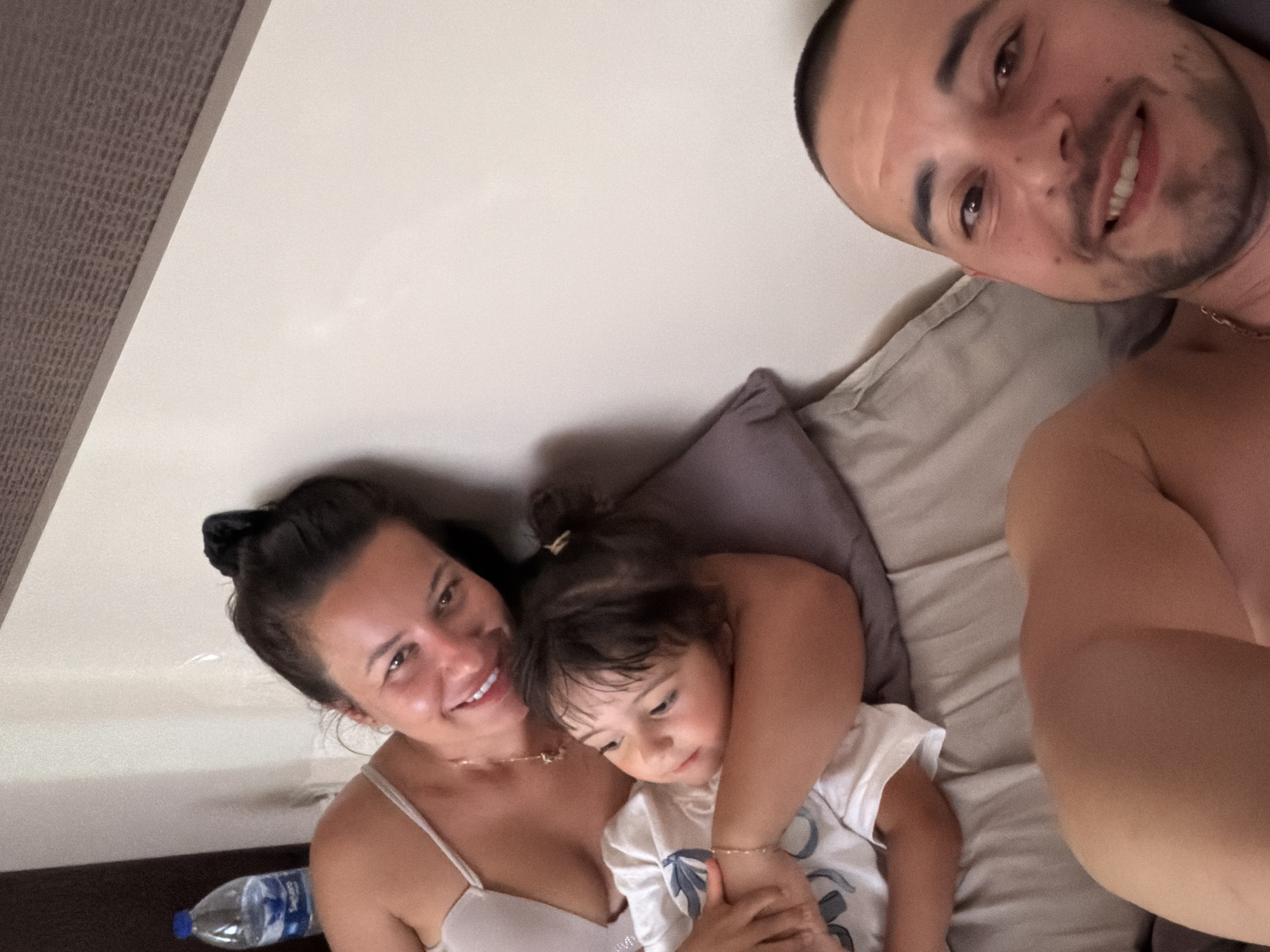 A family is in bed. A woman is lying on her side, with a young child in her arms. A man is lying in bed in front of them, looking at the camera. The scene is indoors, with a light-colored wall and a headboard in the background. There is a bottle of water on a nightstand to the left of the frame. The lighting is soft and natural. The people appear relaxed and happy. The location is a bedroom in a hotel in Burgas, Bulgaria.