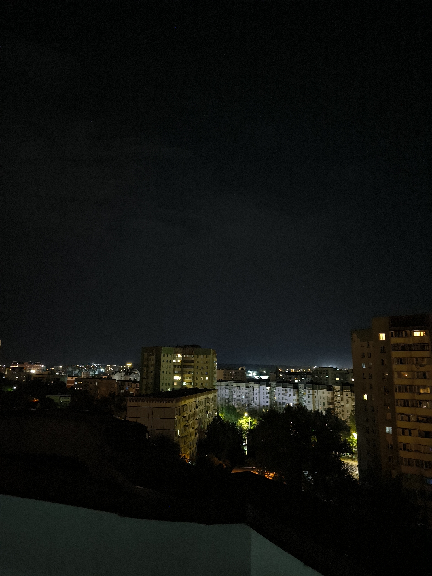 The image shows a nighttime cityscape in Chișinău, Moldova. In the foreground, a dark, angled surface, possibly a balcony or rooftop, occupies the bottom portion of the frame. Beyond this, the scene opens up to reveal a dense urban environment with numerous apartment buildings. The buildings are illuminated from within, with many windows glowing with warm yellow light, indicating that the residents are home. Trees are interspersed among the buildings, their dark silhouettes contrasting with the lit windows. The sky above is dark with a few faint stars visible. The overall impression is of a typical residential area at night in a city.