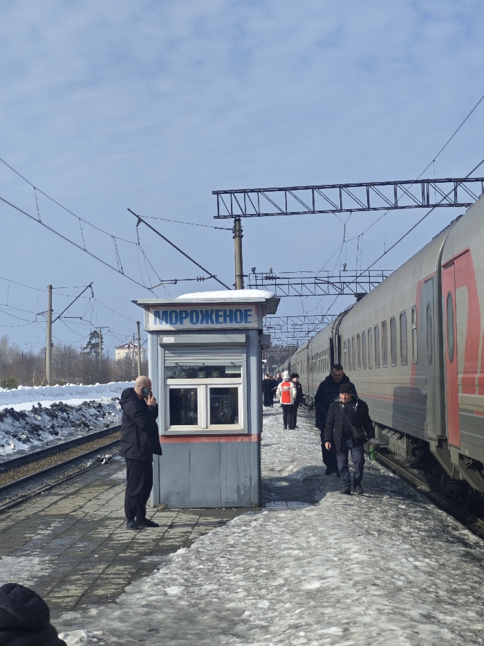 The image captures a train station platform in Druzhinino, Russia, during daylight under a bright, partly cloudy blue sky. A silver-grey passenger train with red accents is stopped at the platform on the right side of the frame. The platform is paved, with visible patches of melting snow and ice. Parallel to the platform, a set of railway tracks runs to the left, bordered by accumulated snow. Overhead electrical lines and supporting utility poles are present above the tracks and platform.
A small, grey kiosk with a white window frame and a closed roller shutter on its front is situated on the platform to the left of the train. The kiosk's roof has a layer of snow, and its sign displays "МОРОЖЕНОЕ" (ICE CREAM) in blue Cyrillic letters. A bald man in dark clothing stands to the left of the kiosk, facing right, holding a dark smartphone to his right ear as if engaged in a call.
On the platform to the right, adjacent to the train, several individuals are visible. Two men in dark jackets and pants walk away from the camera, along the platform next to the train. Further down the platform, a person wearing a red and white jacket stands facing left, with additional indistinct figures visible in the distance. The overall environment suggests a winter or early spring setting, with bare trees and some evergreens in the background.