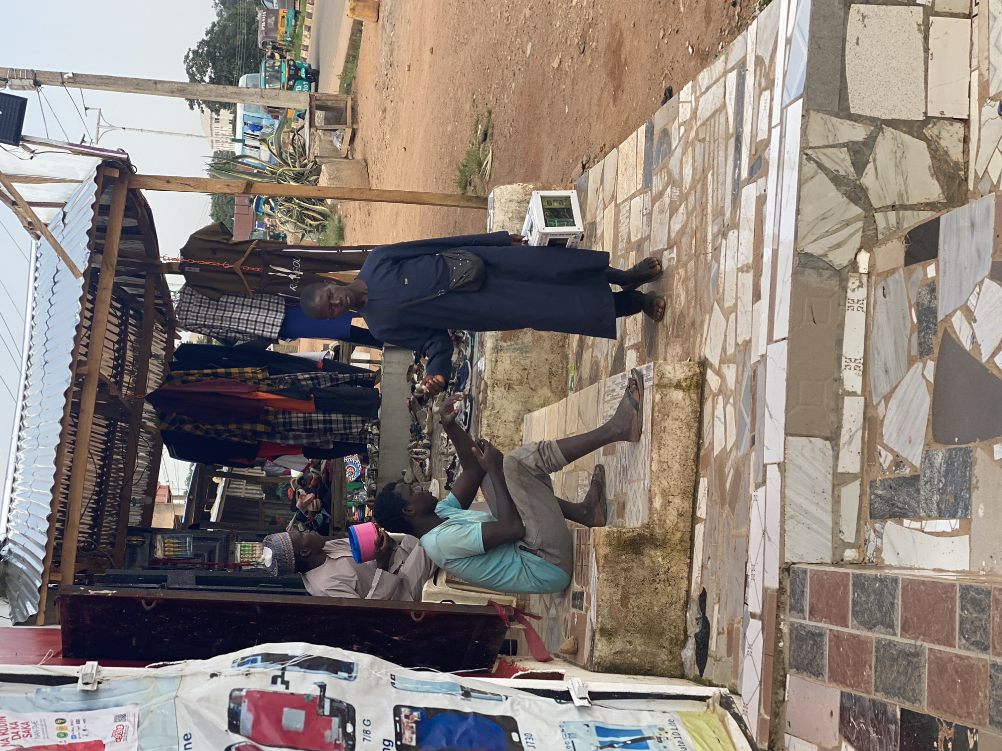 In Gura Top, Nigeria, two men are visible at an outdoor market stall. One man, wearing a dark blue robe and a black crossbody bag, stands facing left. Another man, in a light blue t-shirt and grey pants, is seated, stretching his legs. Behind them, a stall displays various clothing items, including shirts and pants. A smaller stand to the right holds what appears to be a computer tower and some electronic components. The ground is a mix of dirt and mosaic tiles. In the background, a road is visible with several auto-rickshaws, suggesting a busy, daytime scene. The weather appears to be overcast. The visible text includes brand names and product information, such as "NA RUDIN," "DA KA SAKA," "NAN-TAME," "7/8 G," "J730," and "ate 10 Lite."