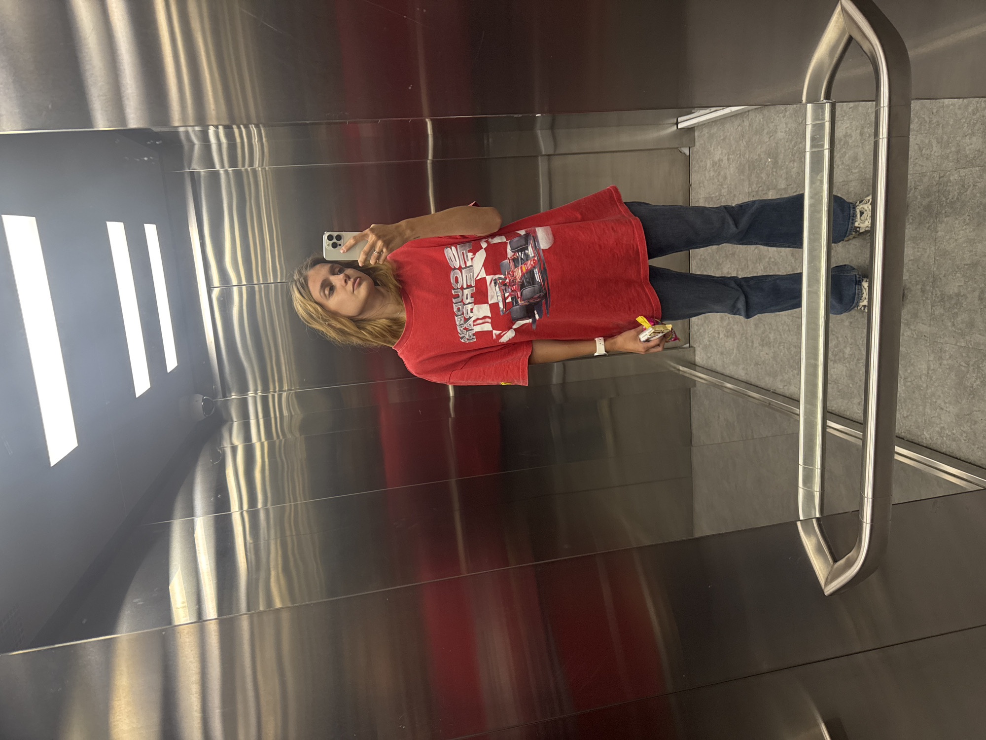 A woman stands inside an elevator, taking a selfie in the mirror. She is wearing a red T-shirt with a race car design, dark jeans, and patterned sneakers. She holds a white smartphone in her right hand and a small yellow object in her left. The elevator has reflective metal walls and floors, and a metal handrail is visible. The environment suggests a modern urban setting. Location: Unknown, Unknown.