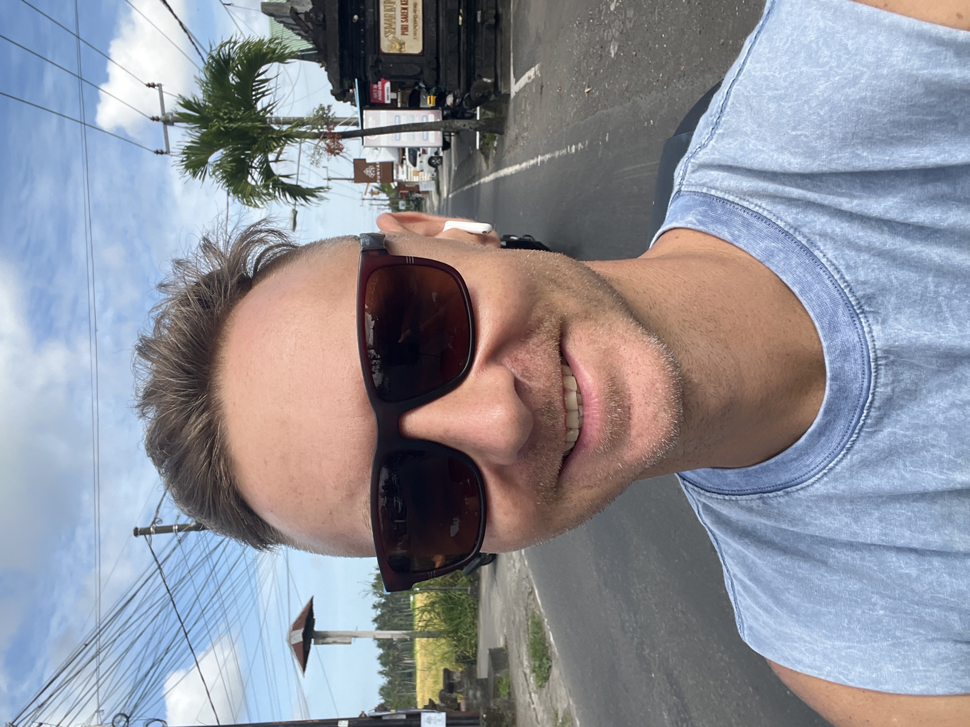 A selfie of a man wearing sunglasses and a blue t-shirt, with an earbud in his left ear, taken on a street in Ubud District, Indonesia. He is on a road with a palm tree to the left and buildings on both sides. Overhead power lines are visible against a blue sky with some clouds. The road appears to be paved and slightly cracked.