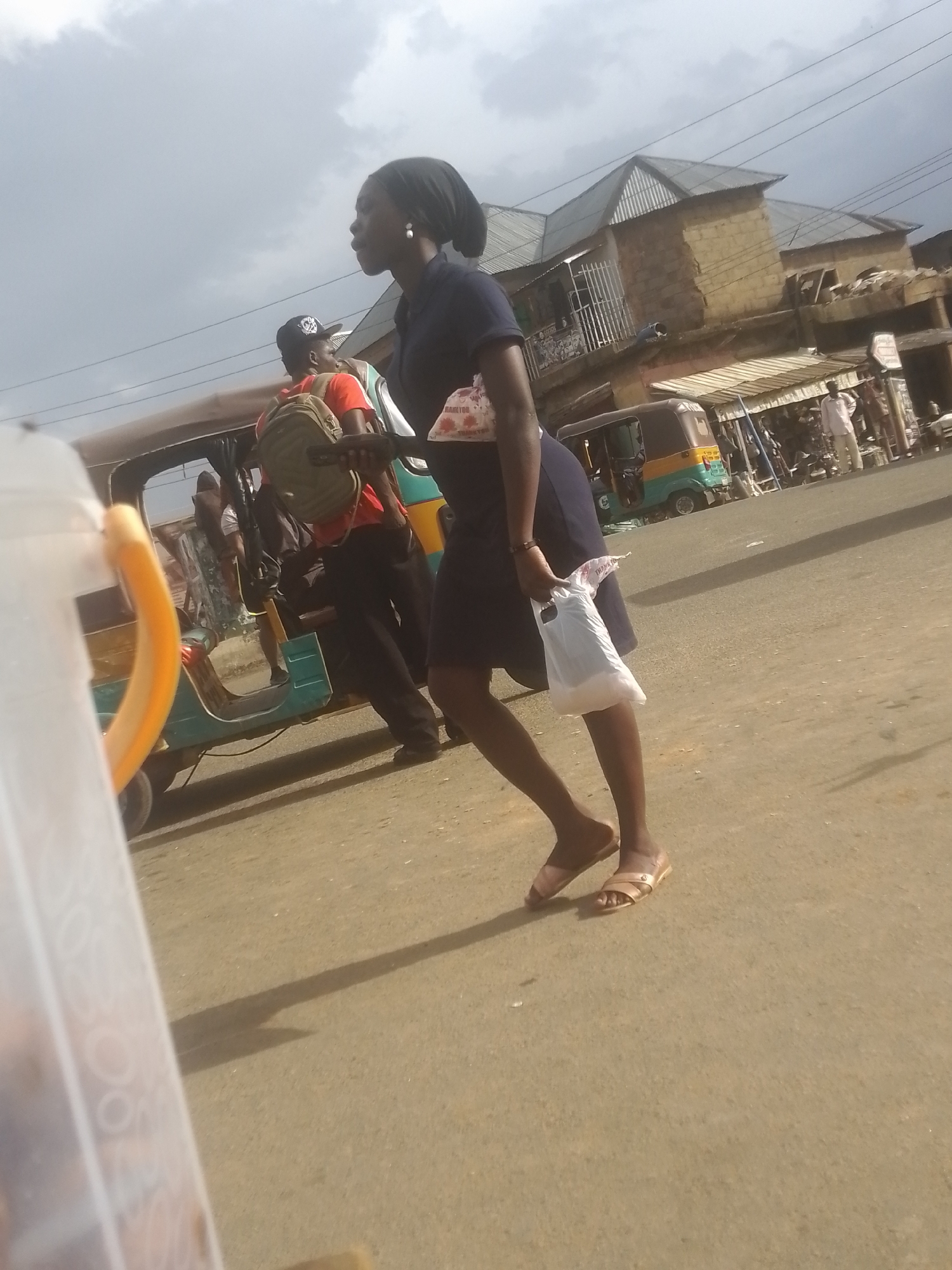 In an outdoor urban setting, a young Black woman walks on a paved surface with a clear sky overhead. She is wearing a dark blue shirt, dark blue skirt, and light brown sandals. The woman has a black headscarf covering her hair and is carrying a white plastic bag in her right hand. In the background, a three-wheeled vehicle, possibly an auto-rickshaw, is parked on the right side of the road. Several buildings and market stalls line the street. Another person wearing a red shirt and a cap can be seen near the auto-rickshaw, with a backpack on. The lighting suggests it is daytime.