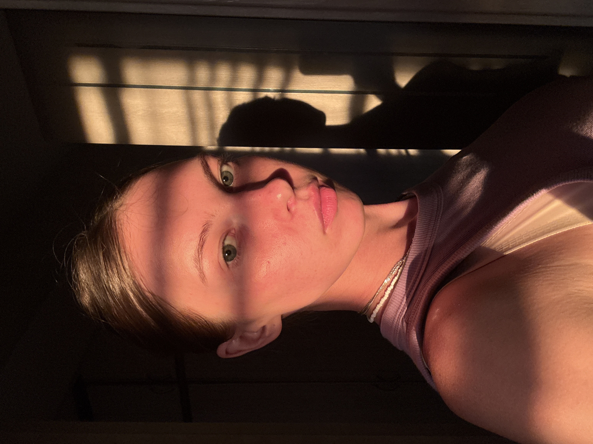 The image presents an indoor, close-up, waist-up portrait of a young adult female, likely a selfie. She has brown hair pulled back, green eyes, and pink lips set in a neutral expression, gazing directly forward. Her skin, particularly on her forehead, left cheek, and nose bridge, shows a reddish-orange hue from a strong, low-angle light source entering from the viewer's left. She is wearing a ribbed, sleeveless, lilac-colored top, beneath which a lighter-colored strap is visible on her right shoulder. Two necklaces are worn: a white beaded strand and a thinner metallic chain. The background to the left is dark and indistinct. To the right, a vertical, light-colored surface, possibly a door or wall, is illuminated, displaying sharply defined shadows. These shadows include vertical stripes, consistent with window blinds or a frame, and the prominent silhouette of a human head and shoulders, which appears to be the subject's own shadow. The scene is situated in an interior location within the city of Perm, Russia.