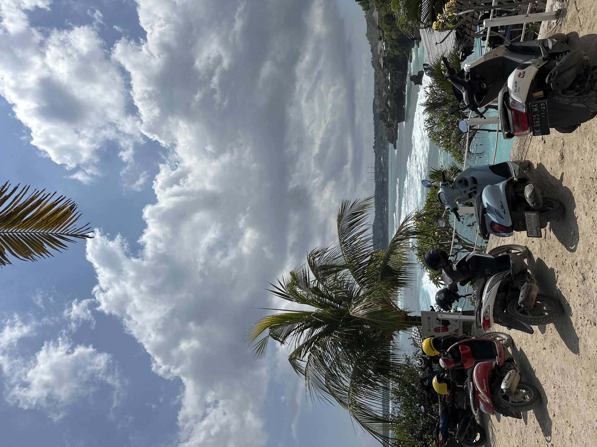 The image captures a scenic viewpoint in Nusa Penida, Indonesia, overlooking a turquoise ocean with crashing waves. In the foreground, a sandy lot is occupied by several parked scooters, their helmets resting on the seats. A large palm tree stands prominently, its fronds casting shadows on the ground. The backdrop features lush green hills dotted with buildings and a hazy coastline. The sky is a mix of blue with scattered white clouds, suggesting a partly sunny day. The overall impression is one of a tranquil, tropical destination.