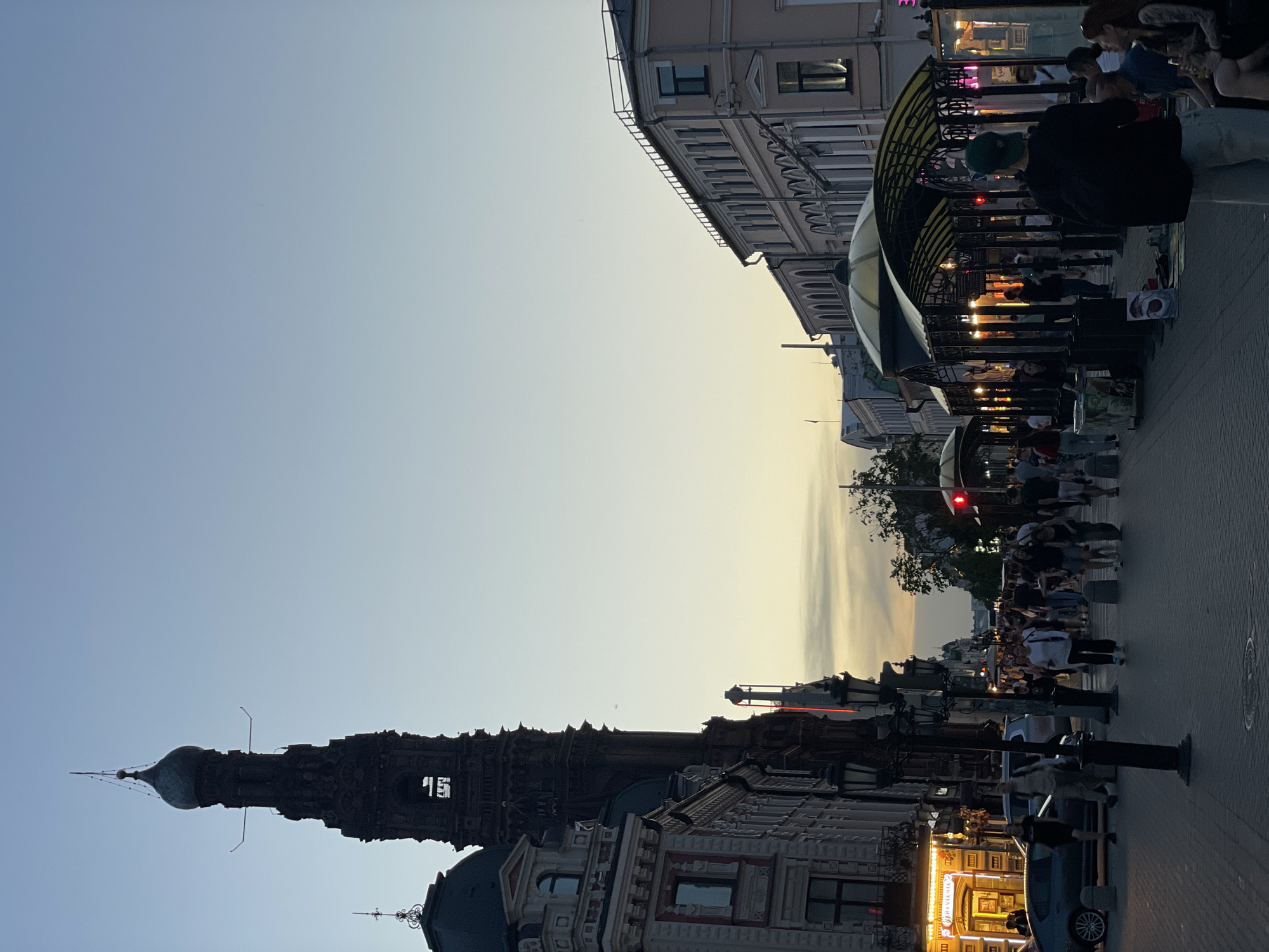 Captured from a low-angle perspective, the scene showcases a bustling pedestrian zone in Kazan, Russia, during the early evening. On the left, the Soyembika Tower stands tall, its digital clock displaying "15." To the right, a line of commercial establishments and cafes line the street, with people populating the sidewalk. The sky transitions from blue to shades of orange as the sun sets.
