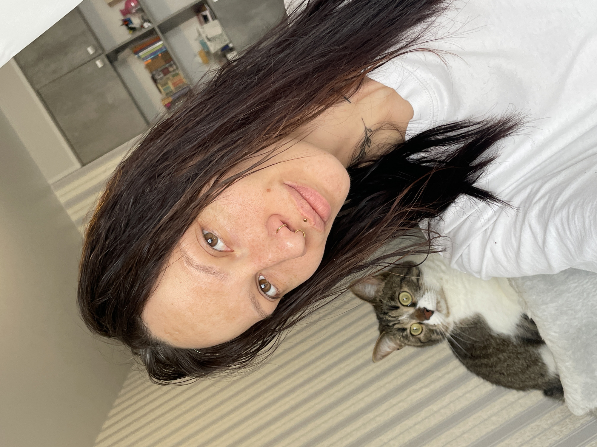 A selfie image presents a woman with brown hair, a nose piercing, and visible freckles. She is positioned on a striped bedspread or sheet, with a white blanket or sheet partially covering her. Adjacent to her, a cat with tabby and white markings is looking up at the camera. In the background, there is a blurred shelving unit with various items. The location is unknown, and there is no visual information in the image to suggest it is Izhevsk, Russia.