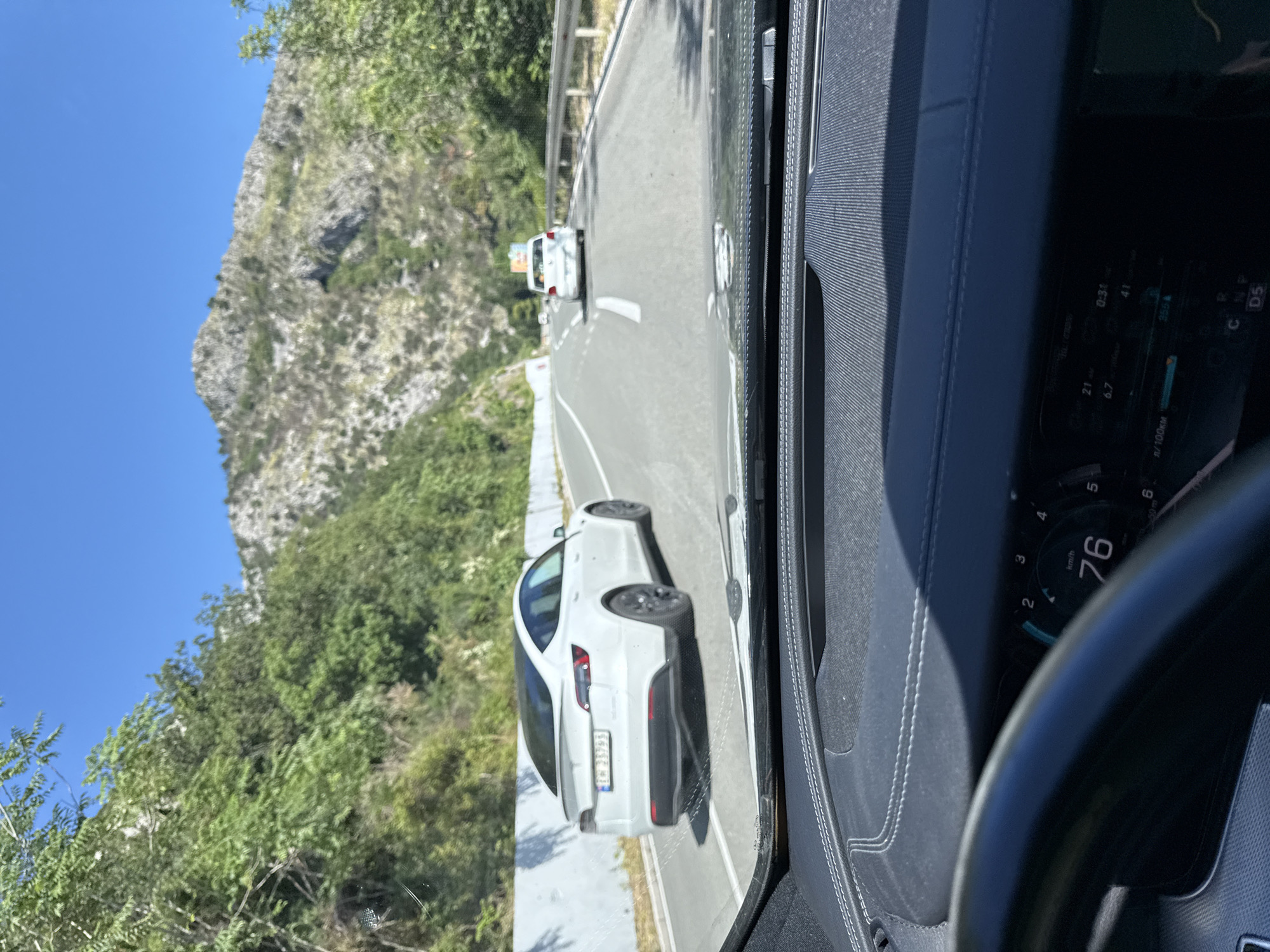 The image captures a view from inside a vehicle, looking forward through the windshield. The vehicle is on a paved road, likely a highway, winding through a mountainous and forested area. In the foreground, the dashboard of the vehicle is visible, with a digital display showing a speed of 76 km/h. A steering wheel is also partially visible on the left. On the road ahead, a white Tesla is driving in the same direction, slightly to the left. Further ahead, another white car is visible in the distance. The road is flanked by a low concrete barrier on the left and a metal guardrail on the right. The landscape is characterized by steep, rocky hills covered with dense green vegetation and trees, under a clear blue sky. The location is identified as Golubovići, Montenegro.