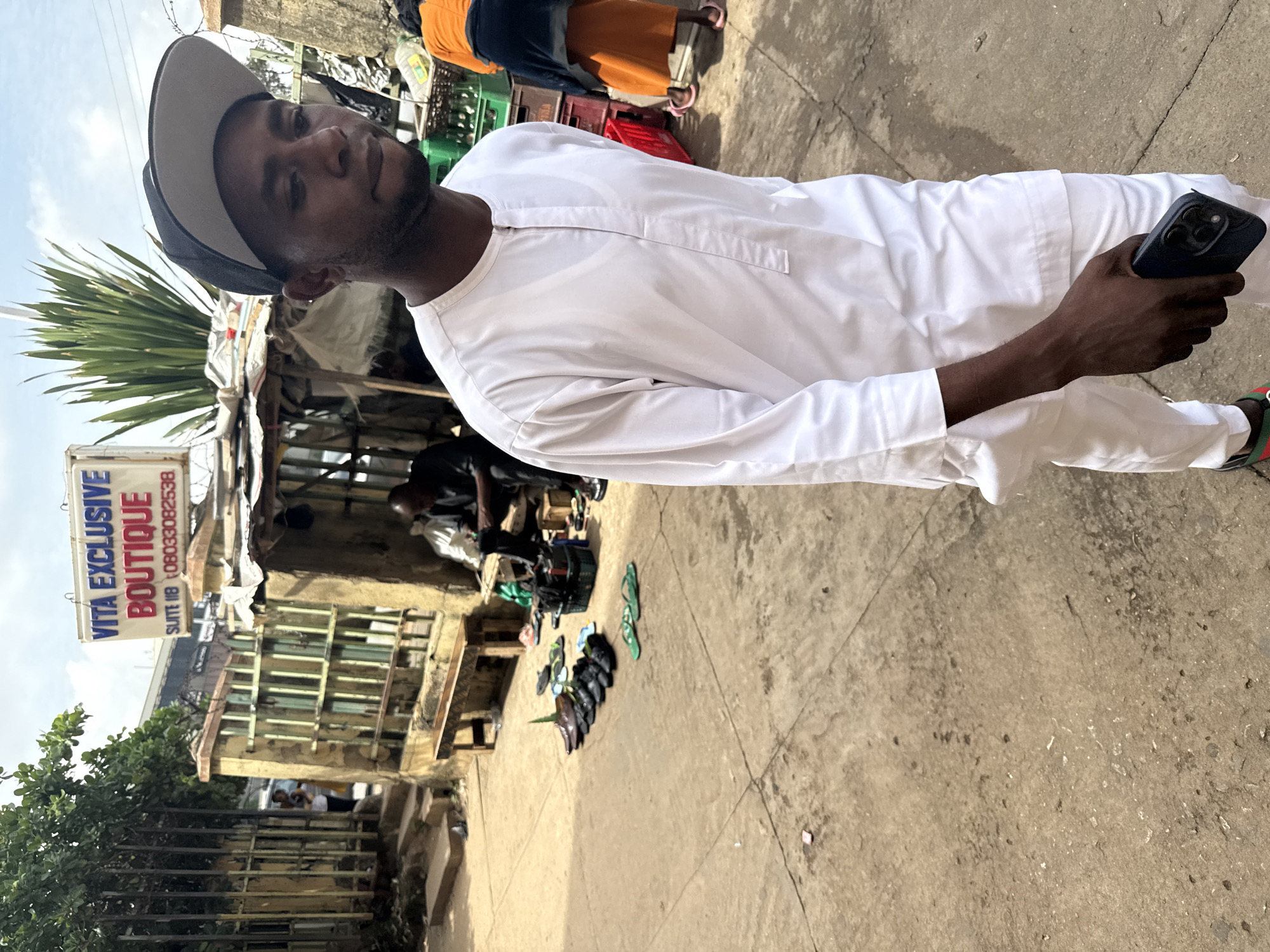 In an outdoor urban setting in Abuja, Nigeria, a man in white traditional attire and a baseball cap walks towards the camera, holding a dark-colored smartphone in his right hand. The background features a modest storefront with a sign reading "VITA EXCLUSIVE BOUTIQUE SUITE 11B 08033082538". To the left, a metal gate with lush green foliage stands beside a weathered stone wall. In front of the boutique, several pairs of men's shoes and sandals are displayed on the ground, suggesting a shoe vendor. Another person, wearing an orange top and skirt, walks away from the camera in the background. The ground is paved with concrete, showing signs of wear and tear.