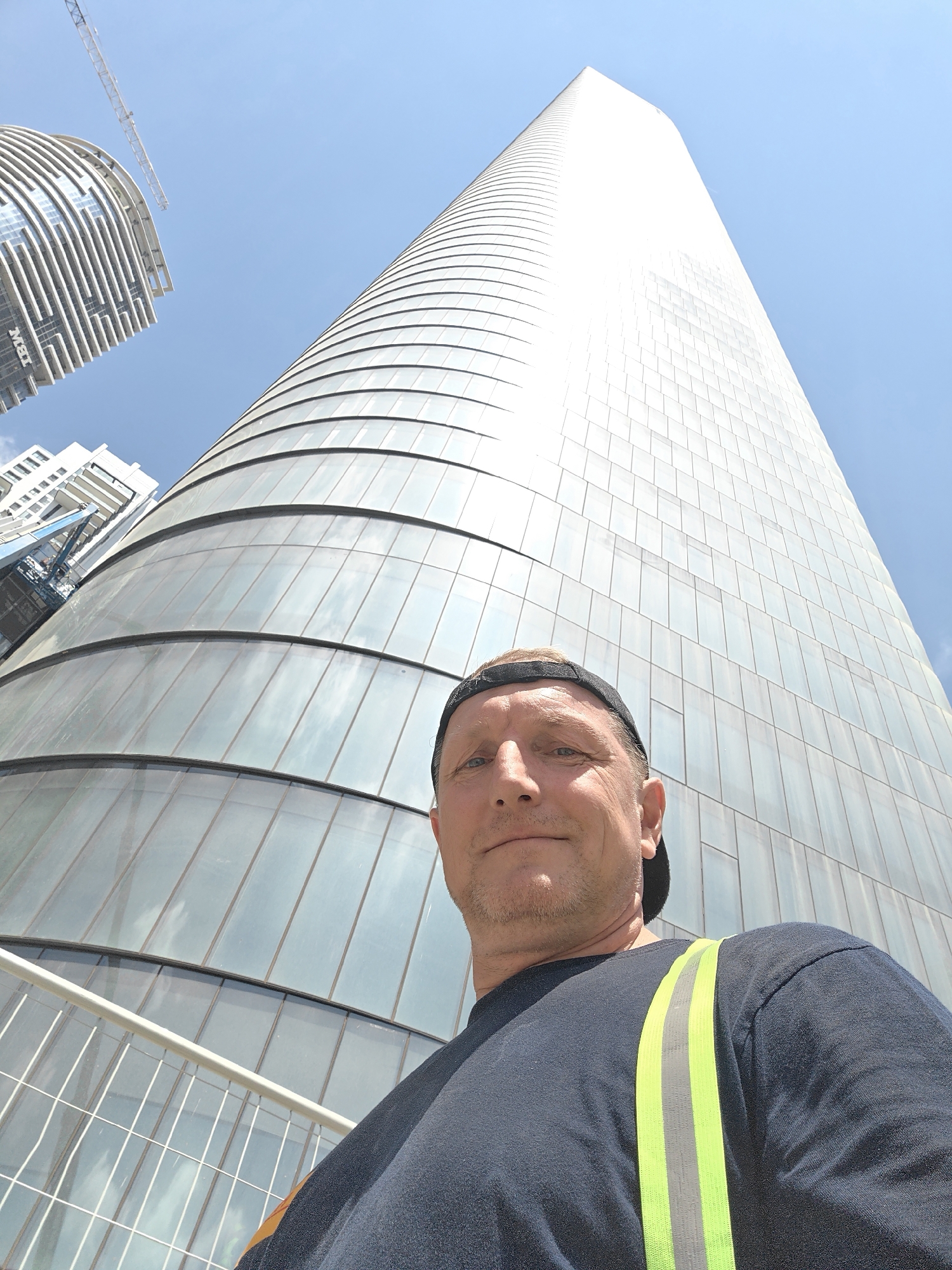 The image shows a man taking a selfie in front of a tall, modern skyscraper with a curved, glass facade. The man is wearing a dark t-shirt with a neon yellow and silver reflective vest over it, and a black baseball cap. He is positioned in the foreground, with the skyscraper dominating the background and filling most of the frame. The sky is clear and blue, with a few clouds visible. To the left of the main skyscraper, another building with a similar architectural style and a construction crane are visible. The perspective is from a low angle, looking up at the structures. The scene appears to be an urban environment. This is the Electra Tower in Givatayim, Israel.