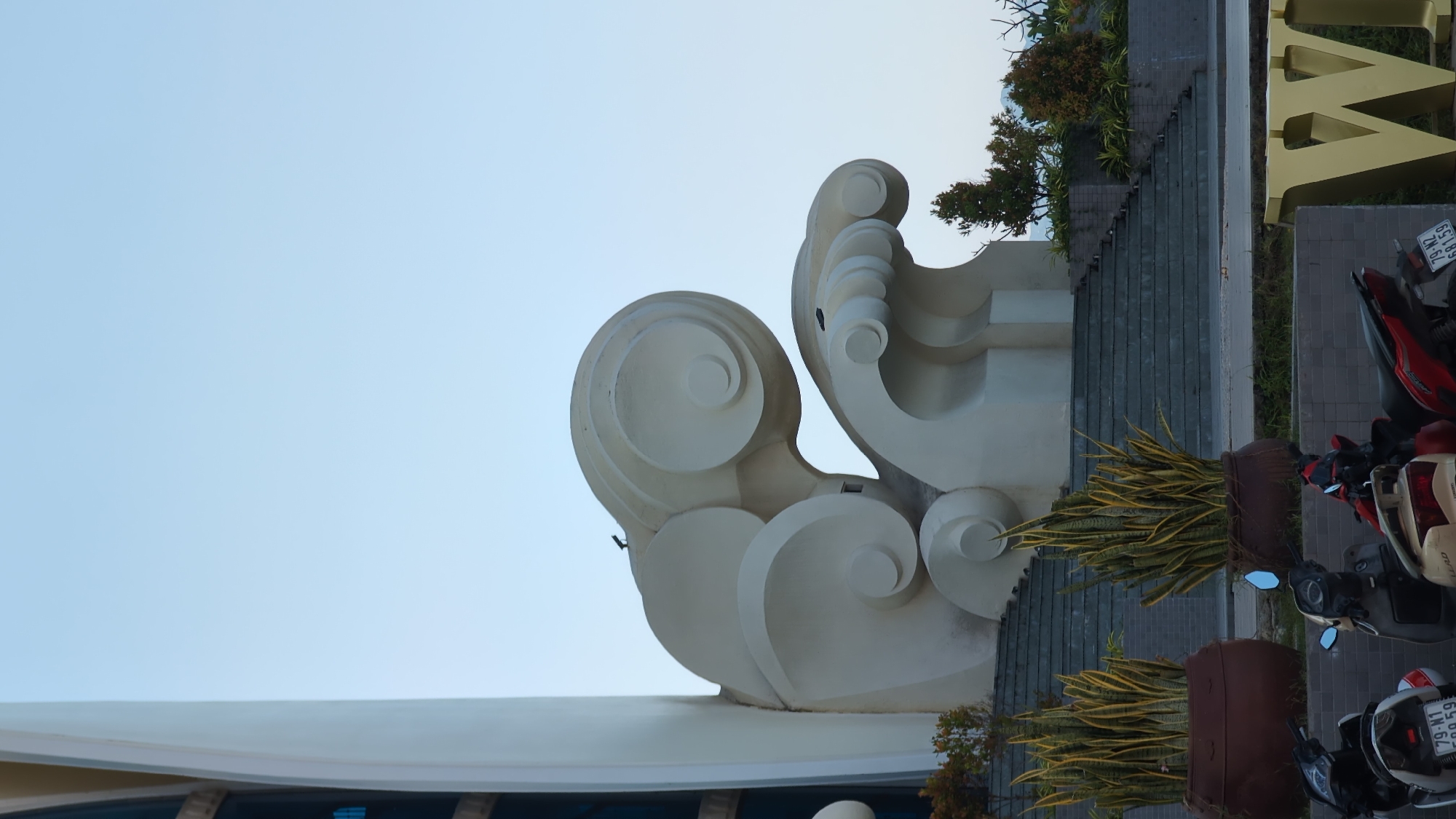 A large, abstract, cream-colored sculpture resembling crashing waves is the central focus of the image, set against a clear, pale blue sky. The sculpture is situated on a tiered stone staircase, with two large, dark brown planters containing spiky, yellow-green plants flanking the lower steps. To the left, a portion of a tall, modern building with dark blue glass windows is visible. In the foreground, several motorcycles are parked. To the right, a large, golden, three-dimensional letter "W" is partially visible. The scene appears to be an outdoor public space, likely in Nha Trang Ward, Vietnam, given the architecture and vegetation. There are no people visible in the image, and the focus is on the art installation and its surroundings. Two small dark birds are perched on the sculpture, one on the top wave and another on the lower wave crest. The overall lighting suggests it is either early morning or late afternoon.