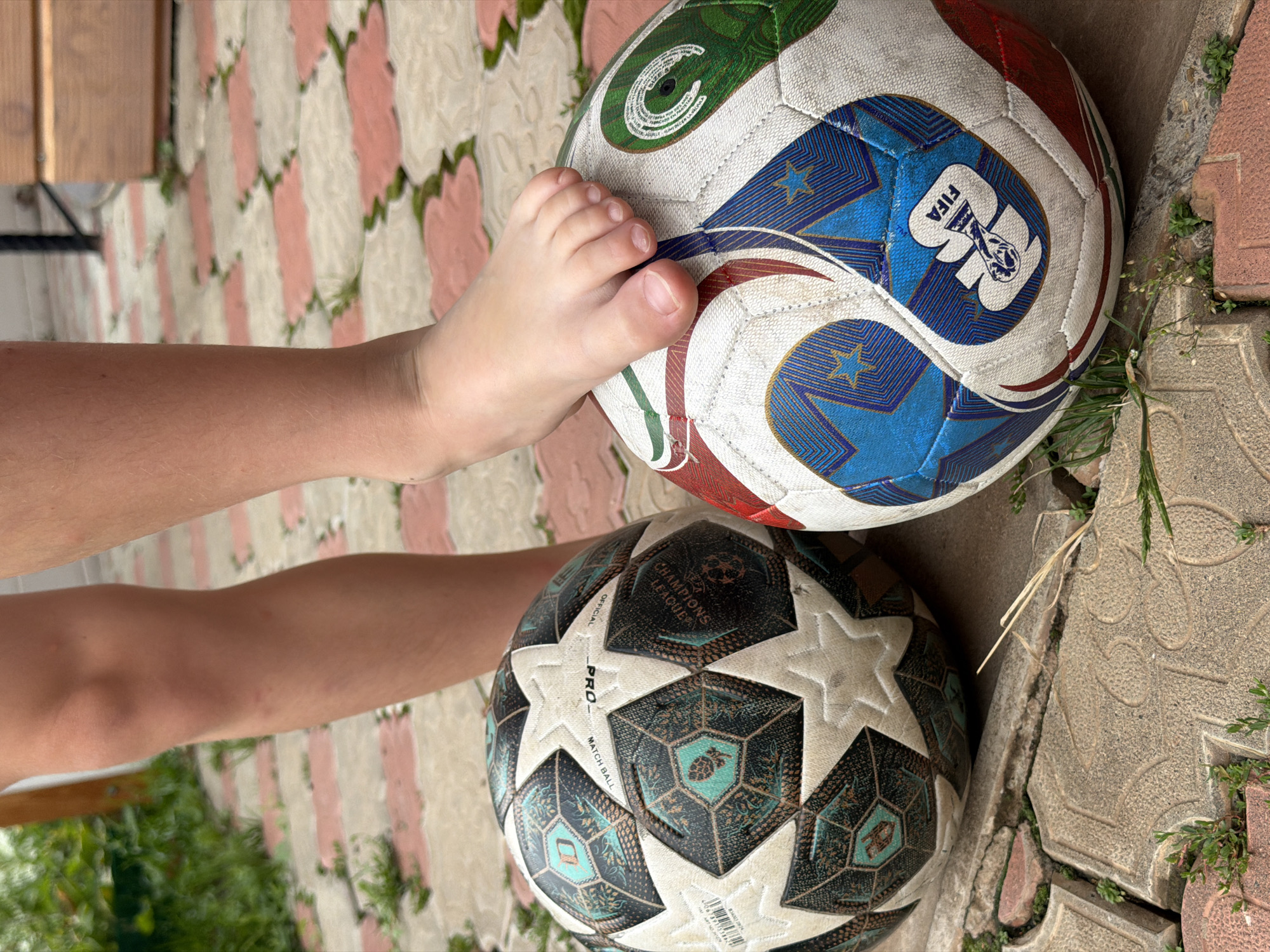 An outdoor, medium-shot photograph shows a child's bare foot resting on a white, blue, and red soccer ball. To the left of this ball is another soccer ball, predominantly white and black with a teal design, which appears to be older and more worn. Both balls are positioned on a stone-tiled patio in a yard. The child's legs and knees are visible above the balls, suggesting they are standing. The background includes blurred green foliage and a wooden structure, indicative of a residential outdoor setting. The location is identified as Tenishevo, Russia.