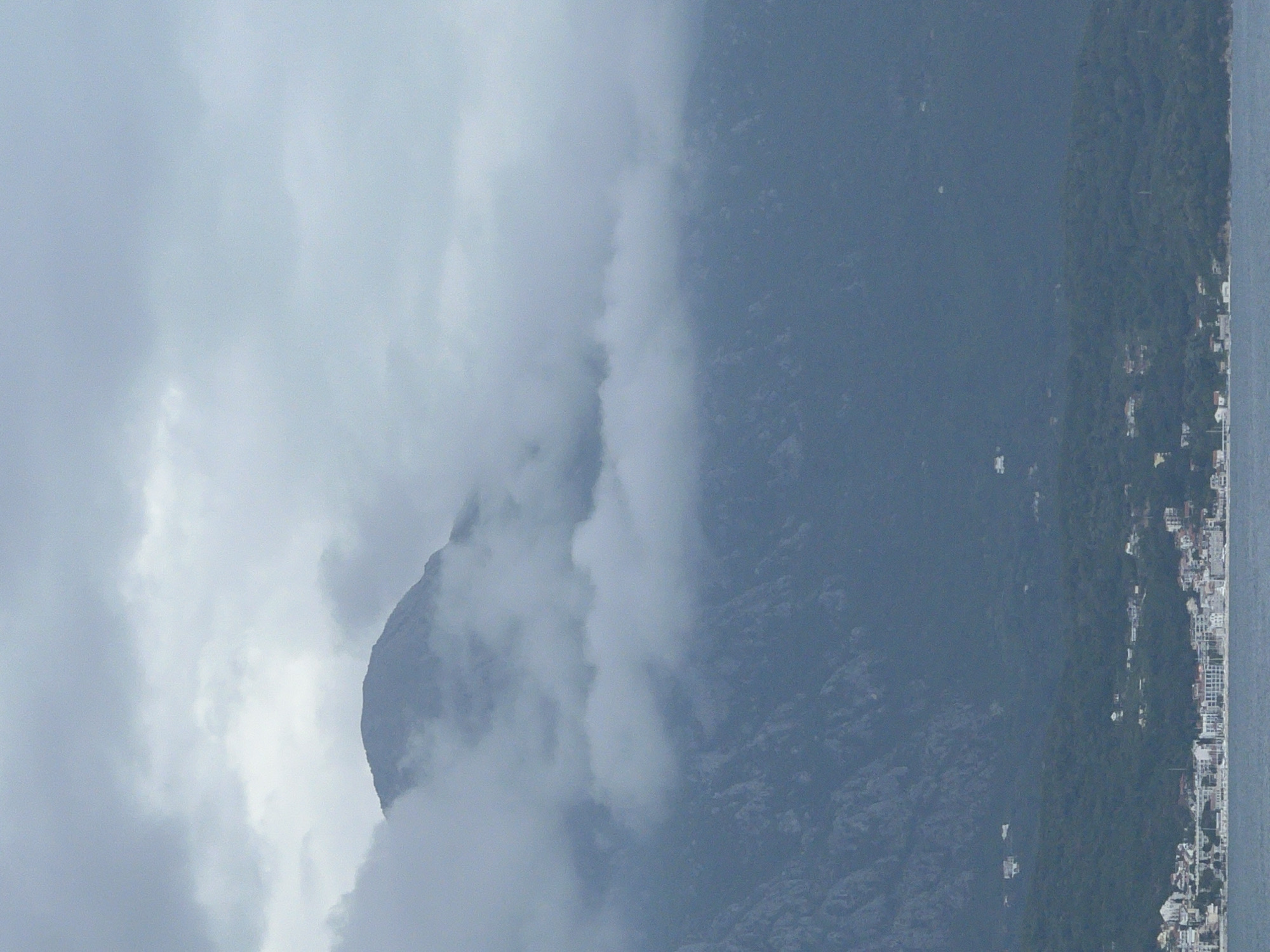 The image shows a mountain partially obscured by clouds. It appears to be located in Luštica, Montenegro, as a town is visible along the shoreline to the right. The mountain is rugged, with visible rock formations. The sky is overcast and gray. The environment appears tranquil and natural.